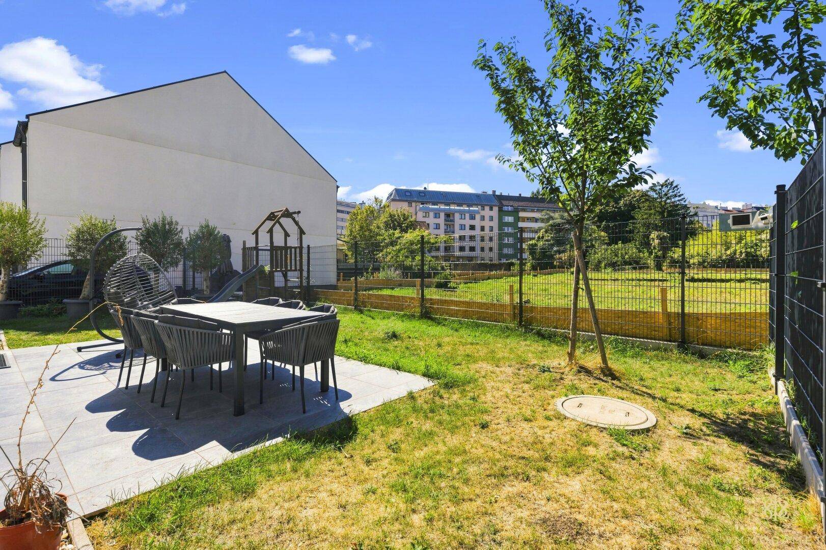 Sonniger Gartenbereich mit gepflasterter Terrasse, Sitzgelegenheiten und Rasenfläche.