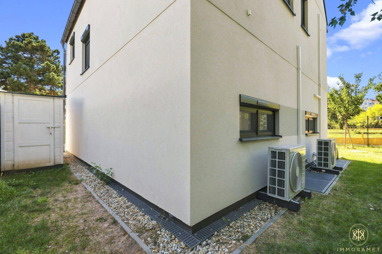 Seitenansicht des Hauses mit Wärmepumpen und einem kleinen Gartenschuppen.