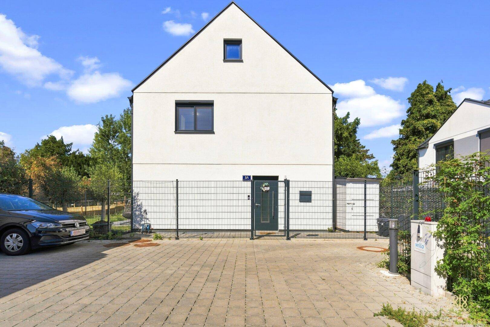 Moderne Hausfassade mit weißem Putz, dunklen Fenstern und gepflastertem Vorplatz.