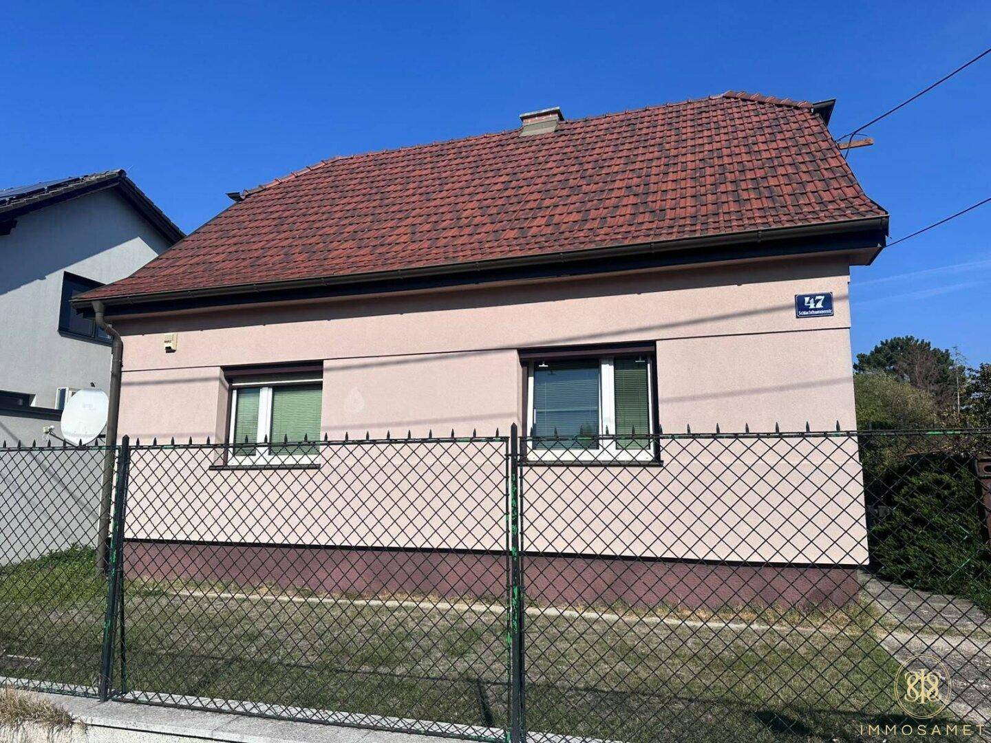 Vorderansicht des Hauses mit rosafarbener Fassade, zwei Fenstern und einem einfachen Zaun.