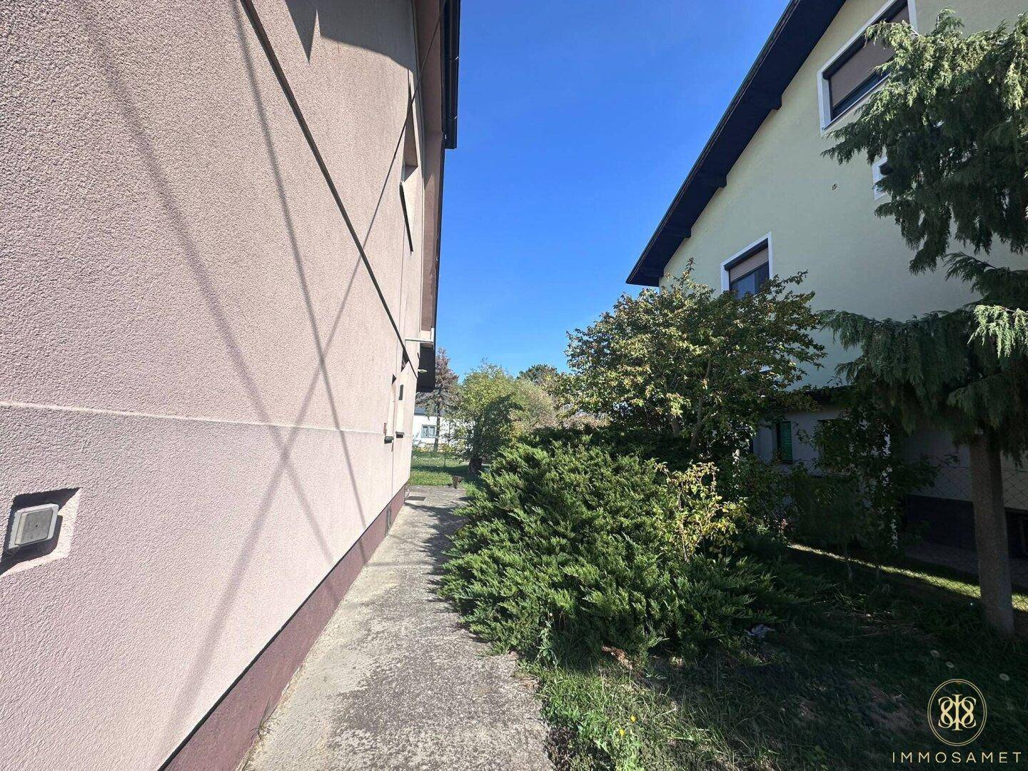Schmaler Weg zwischen zwei Häusern, umgeben von grünen Büschen und Sträuchern.
