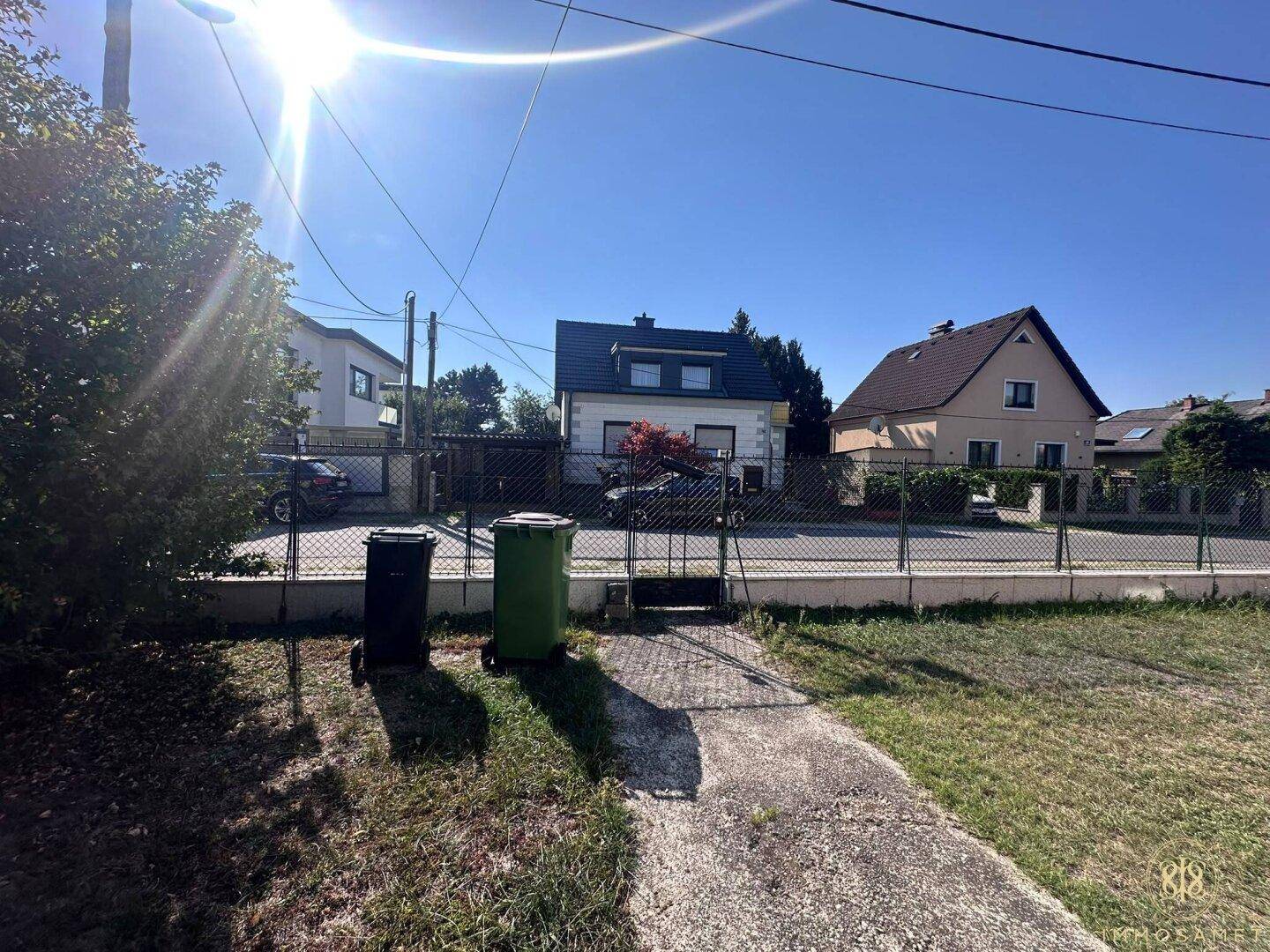 Blick auf die Straße mit weiteren Wohnhäusern und dem Vorgarten mit Mülltonnen.