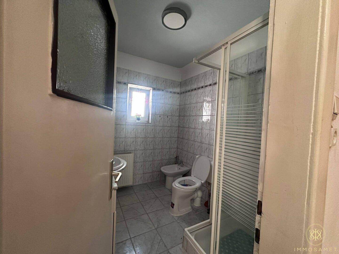 Gefliestes Badezimmer mit Fenster, Duschkabine, WC und Bidet, bietet ausreichend Tageslicht.