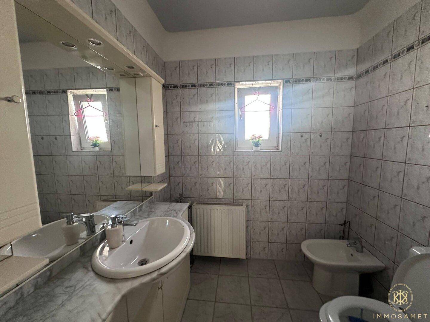 Badezimmer mit zwei Fenstern, Waschbecken, Bidet und WC, in einem traditionellen Stil gehalten.