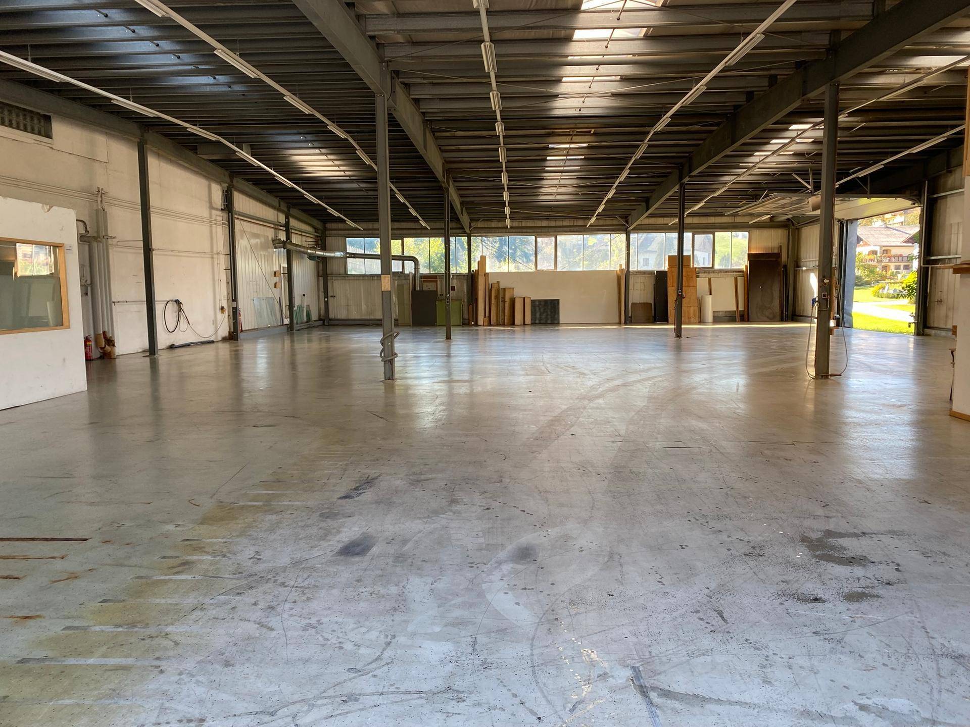 Helle Lagerhalle mit großen Fenstern entlang einer Wand, Betonboden und industrieller Deckenkonstruktion.