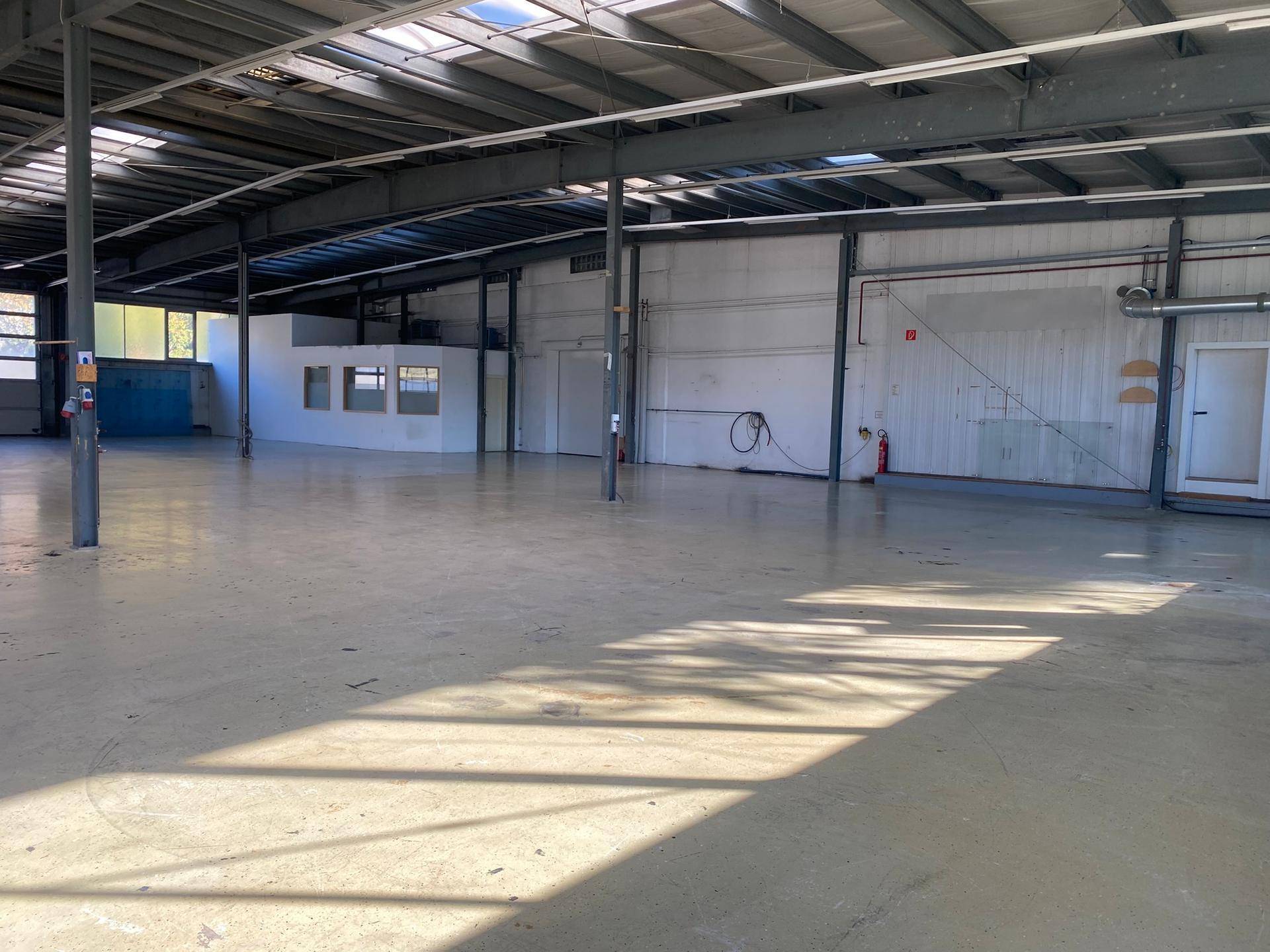 Geräumige, leere Lagerhalle mit Betonboden, Metallträgern und Oberlichtern, gut beleuchtet.