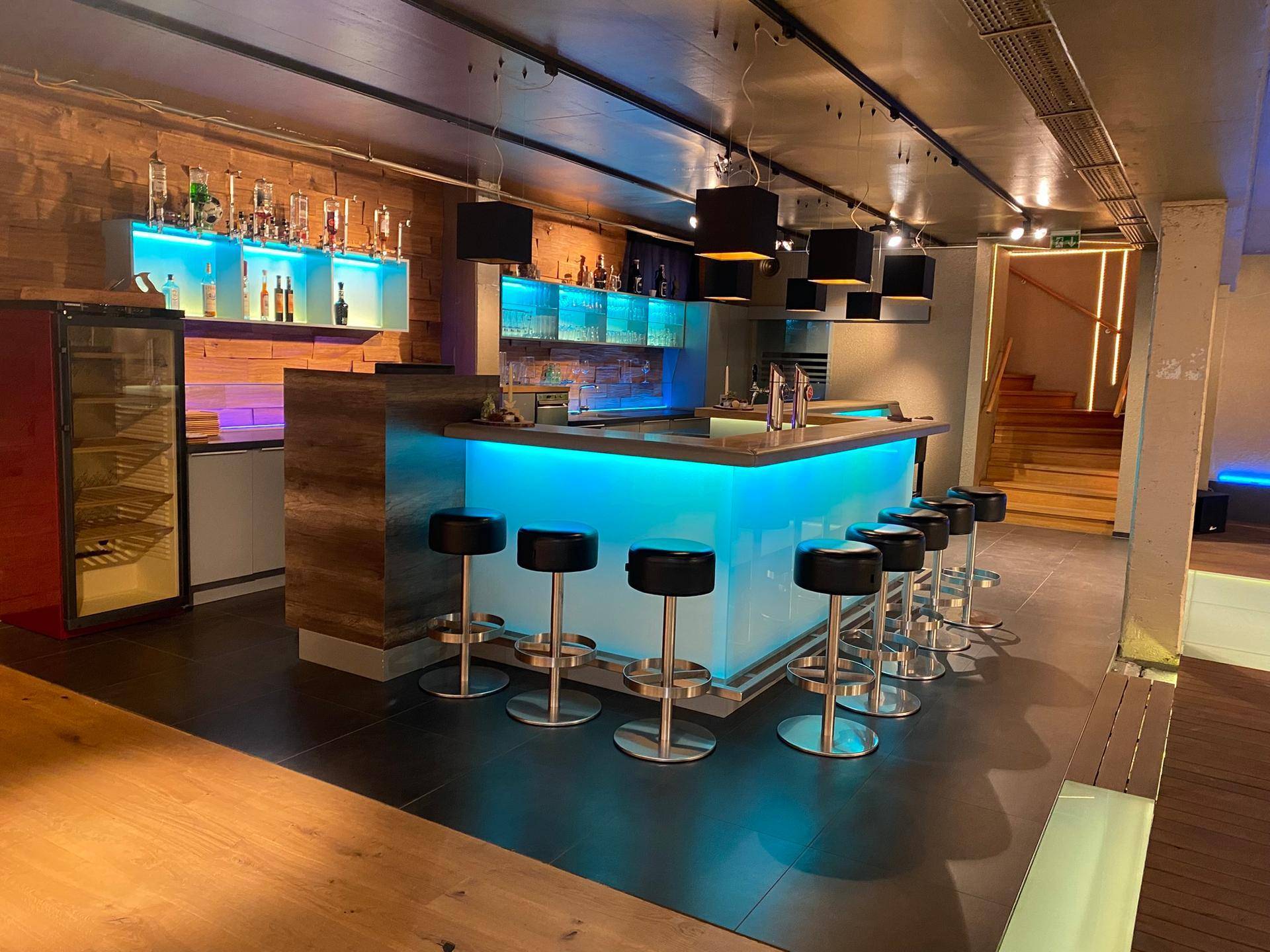 Moderne, beleuchtete Bar mit Barhockern, Flaschenregalen und einem Weinkühlschrank, einladend gestaltet.