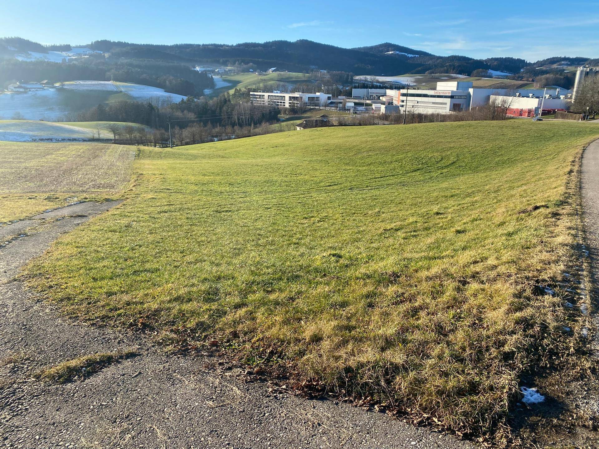 Weitläufiges grünes Feld mit einem Weg, umgeben von sanften Hügeln und Gebäuden in der Ferne.