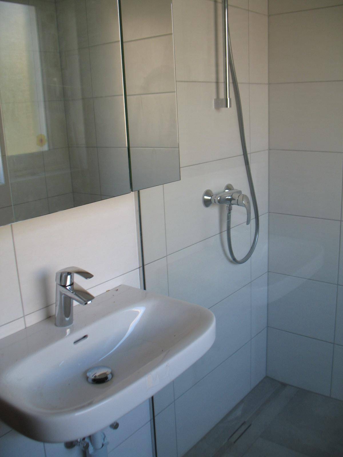 Modernisiertes Badezimmer mit weißem Waschbecken, Spiegel und Duschbereich, hell gefliest.