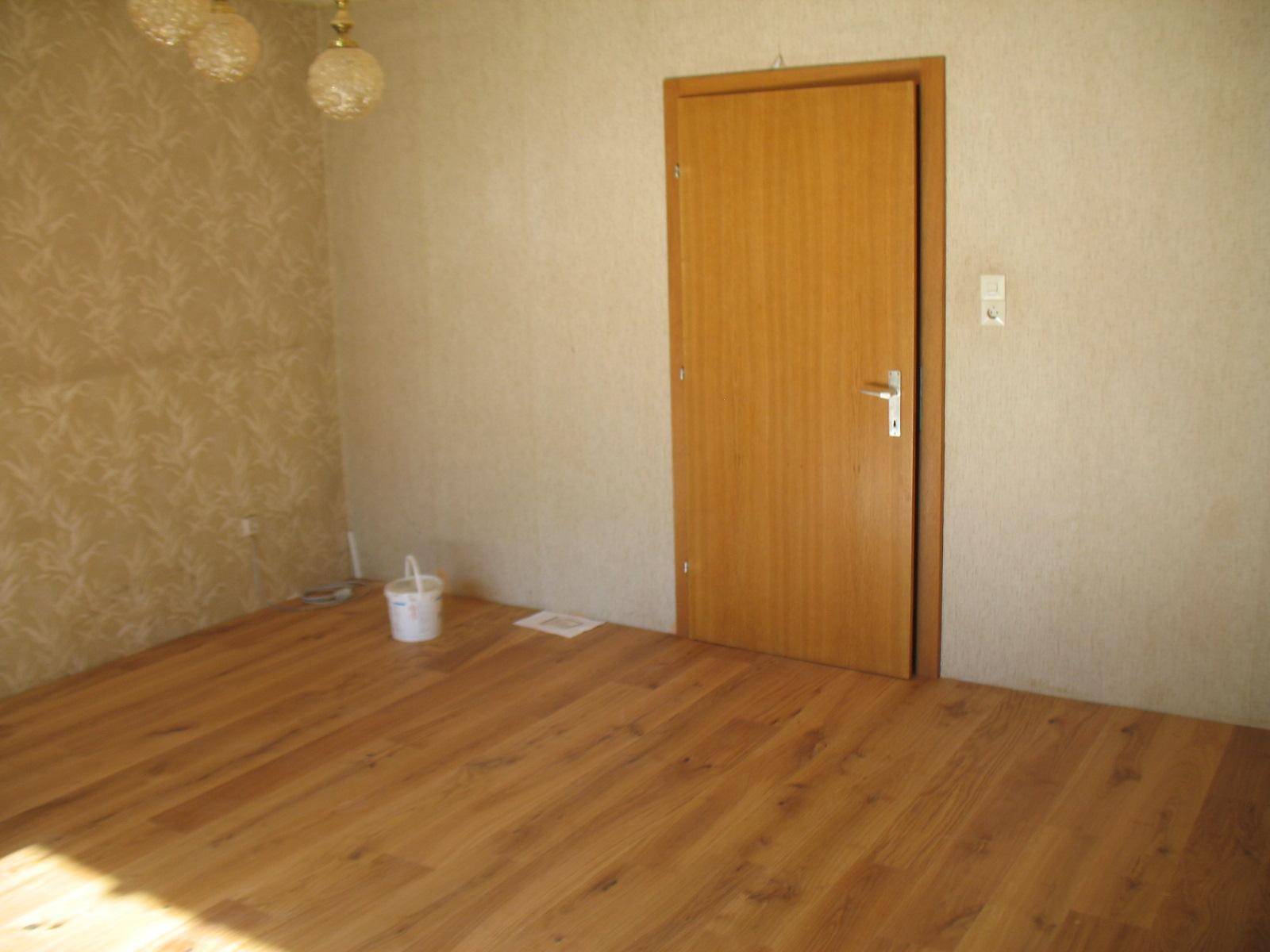 Zimmer mit Holzboden, heller Tapete und einer Holztür, das Renovierungsbedarf aufweist.
