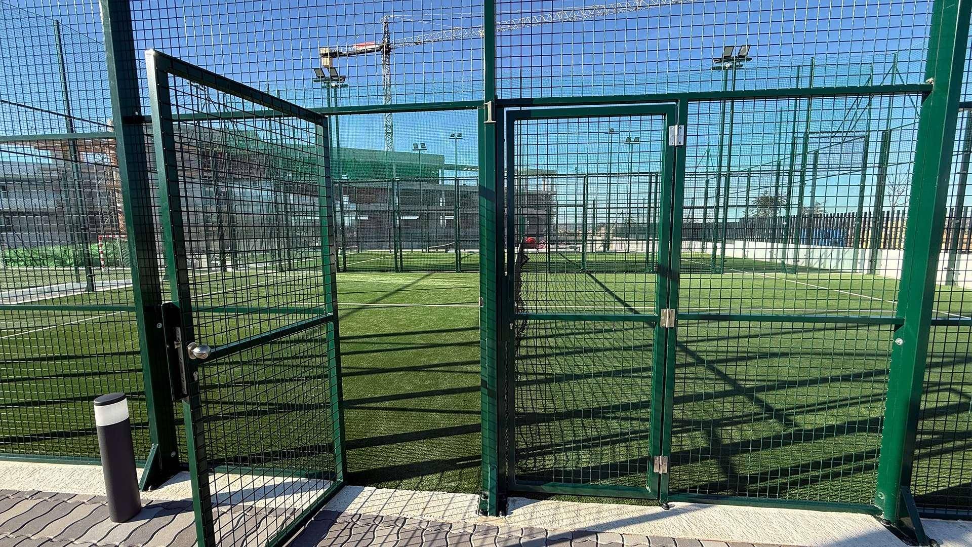 Grüner Padel-Tennisplatz mit Zaun und offenem Tor unter blauem Himmel.