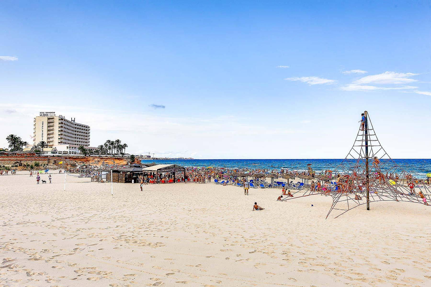 Breiter Sandstrand mit großem Klettergerüst, vielen Badegästen und einem Hotel im Hintergrund.