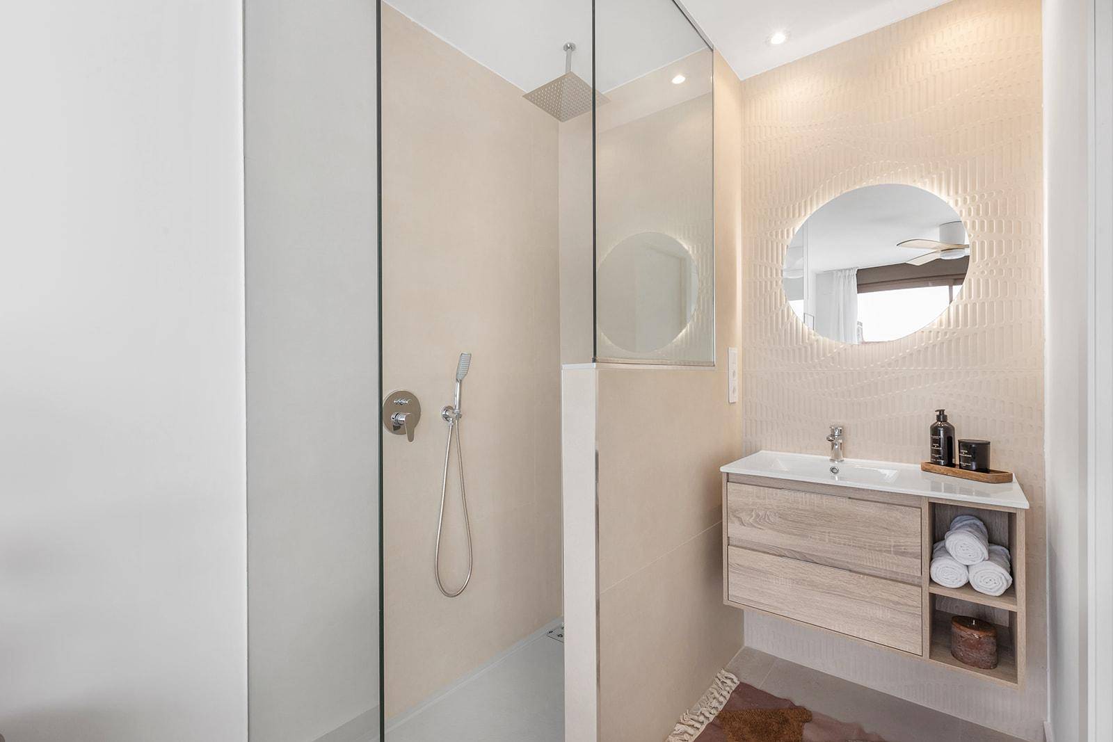 Elegantes Badezimmer mit begehbarer Dusche, runder Spiegelleuchte und Holzunterschrank.