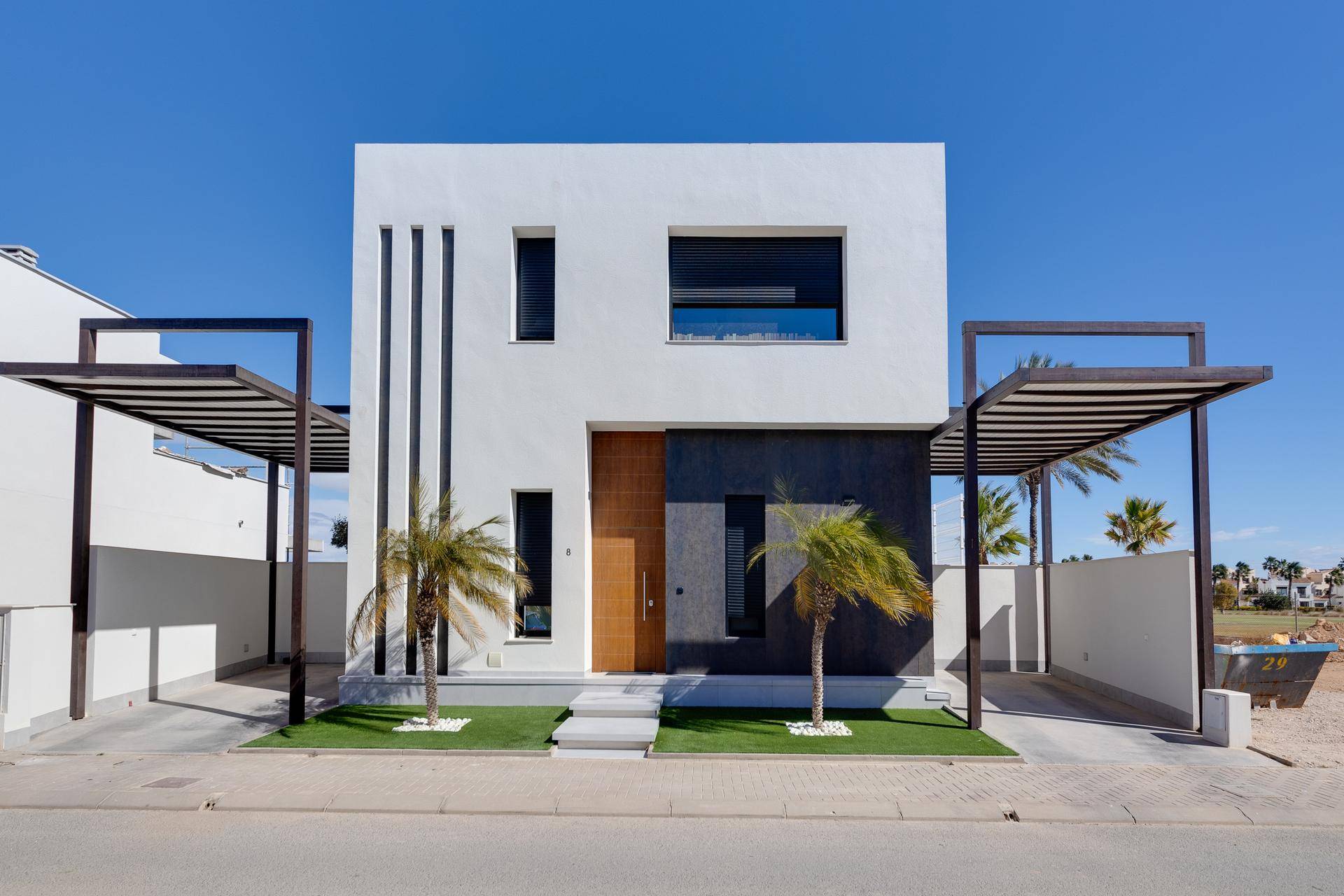 Moderne Hausfassade mit klaren Linien, Holztür und überdachten Parkplätzen unter blauem Himmel.