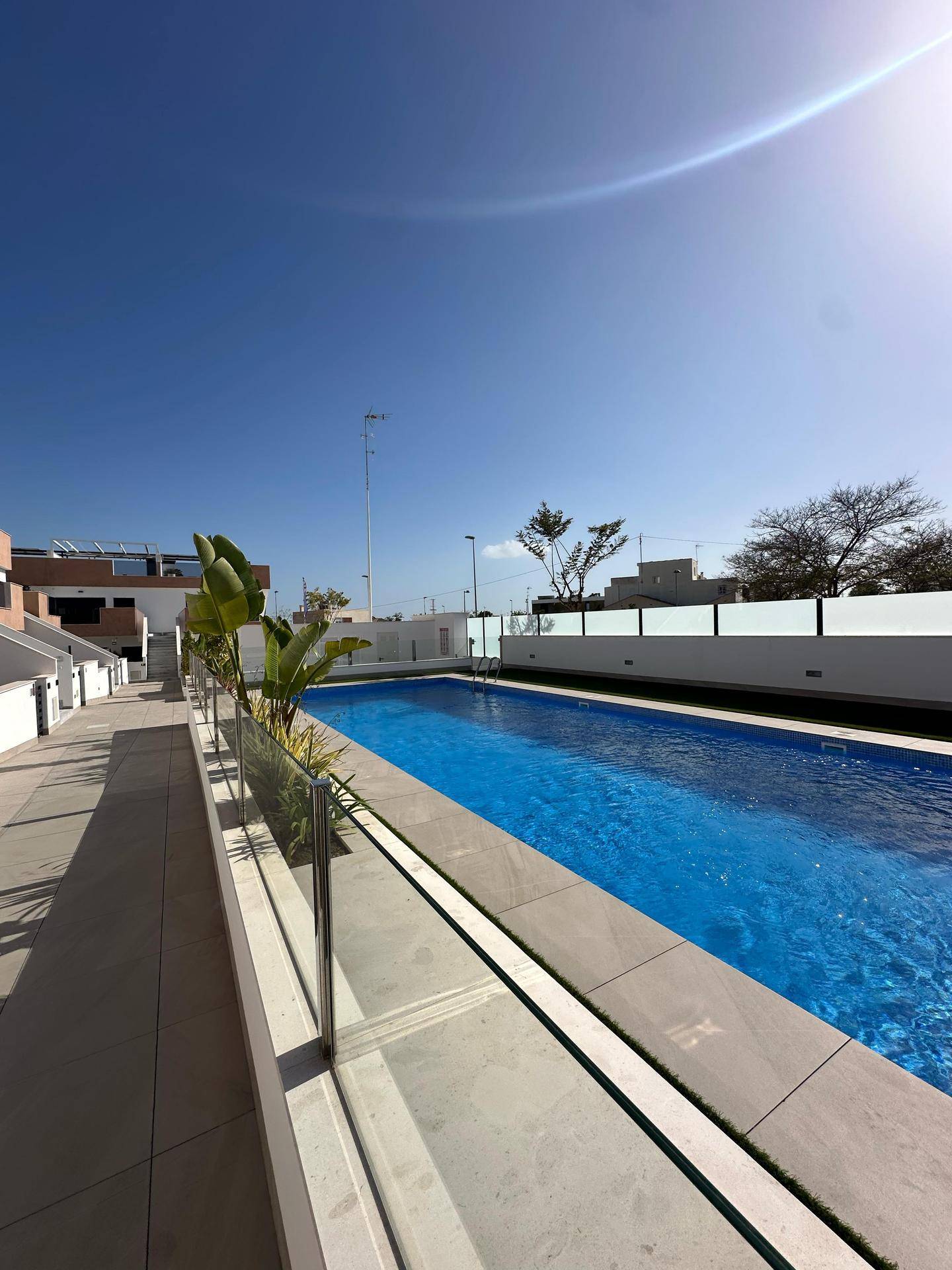Langer Gehweg entlang des Pools mit Glasgeländer und Blick auf die modernen Gebäude.