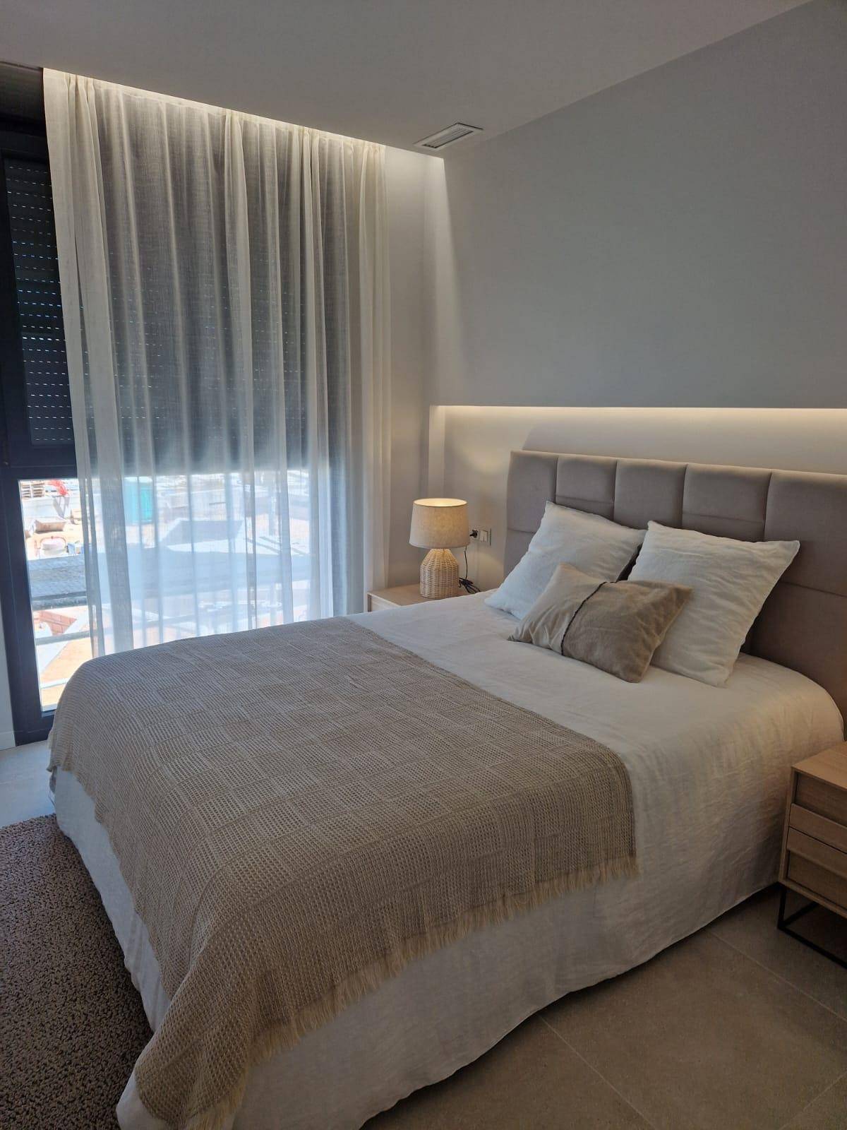 Helles Schlafzimmer mit Doppelbett, Nachttischen und großem Fenster mit transparenten Vorhängen.