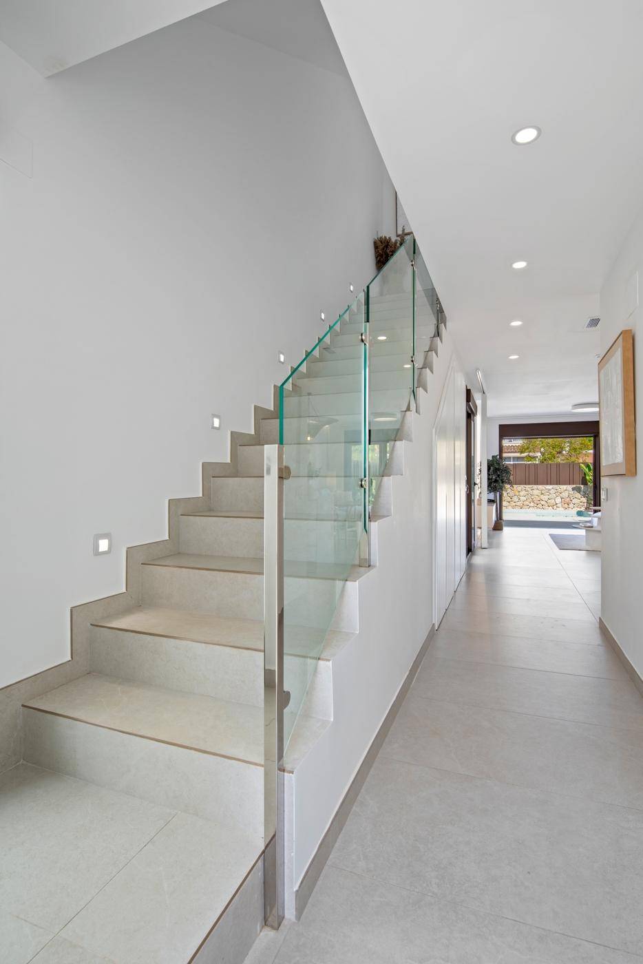 Heller Flur mit moderner Treppe und Glasgeländer, beleuchtet durch integrierte Wandleuchten.