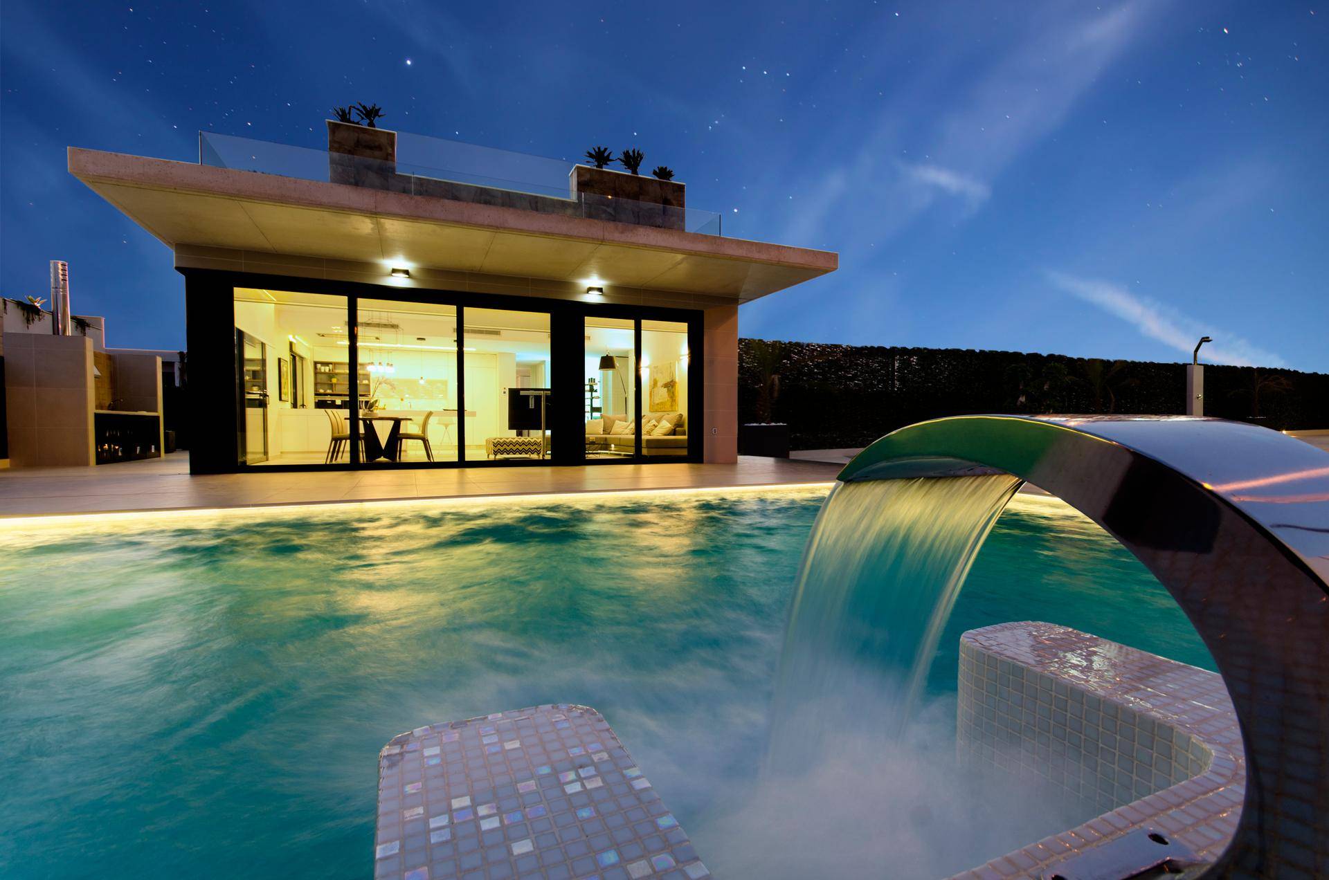 Beeindruckende Villa mit beleuchtetem Pool und Wasserspiel bei Nacht unter sternenklarem Himmel.