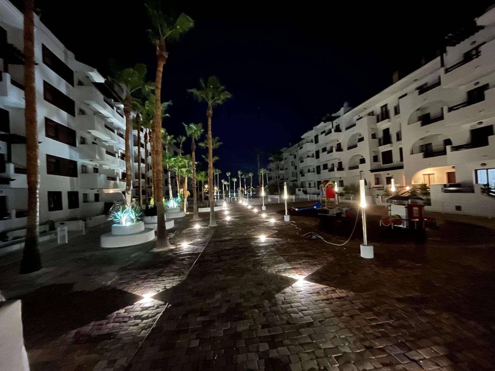 Nachtansicht eines beleuchteten Weges zwischen Apartmentgebäuden, gesäumt von Palmen.