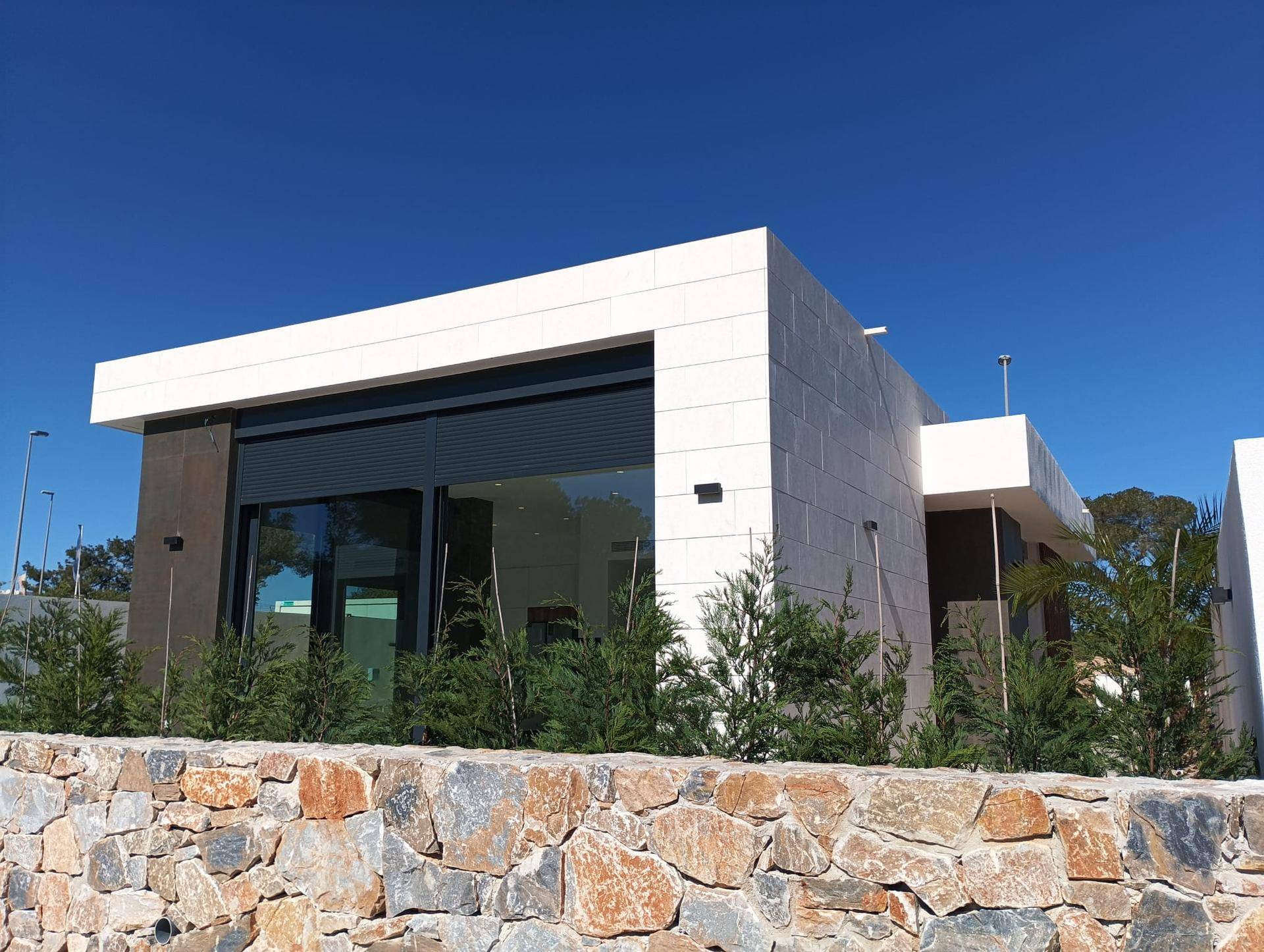 Moderne Architektur der Villa mit weißer Fassade, großen Fenstern und dunklen Rollläden unter blauem Himmel.