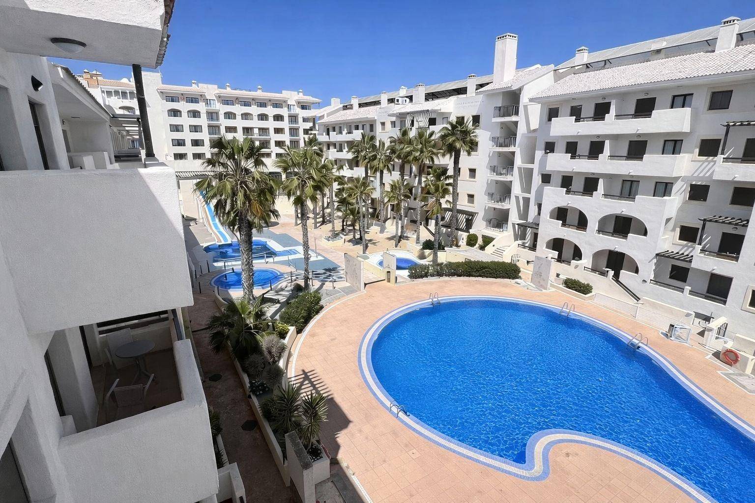 Großer Gemeinschaftspool umgeben von Apartmentgebäuden und Palmen in einer sonnigen Anlage.