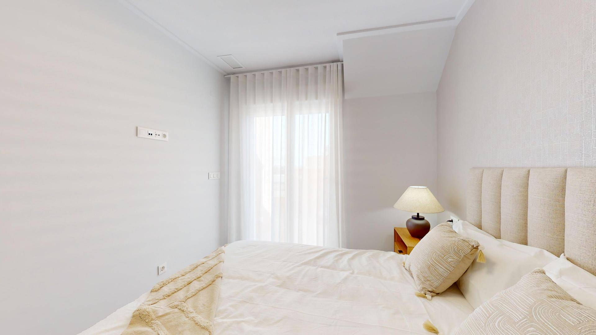 Helles Schlafzimmer mit beigem Kopfteil, weißen Vorhängen und einer Nachttischlampe.