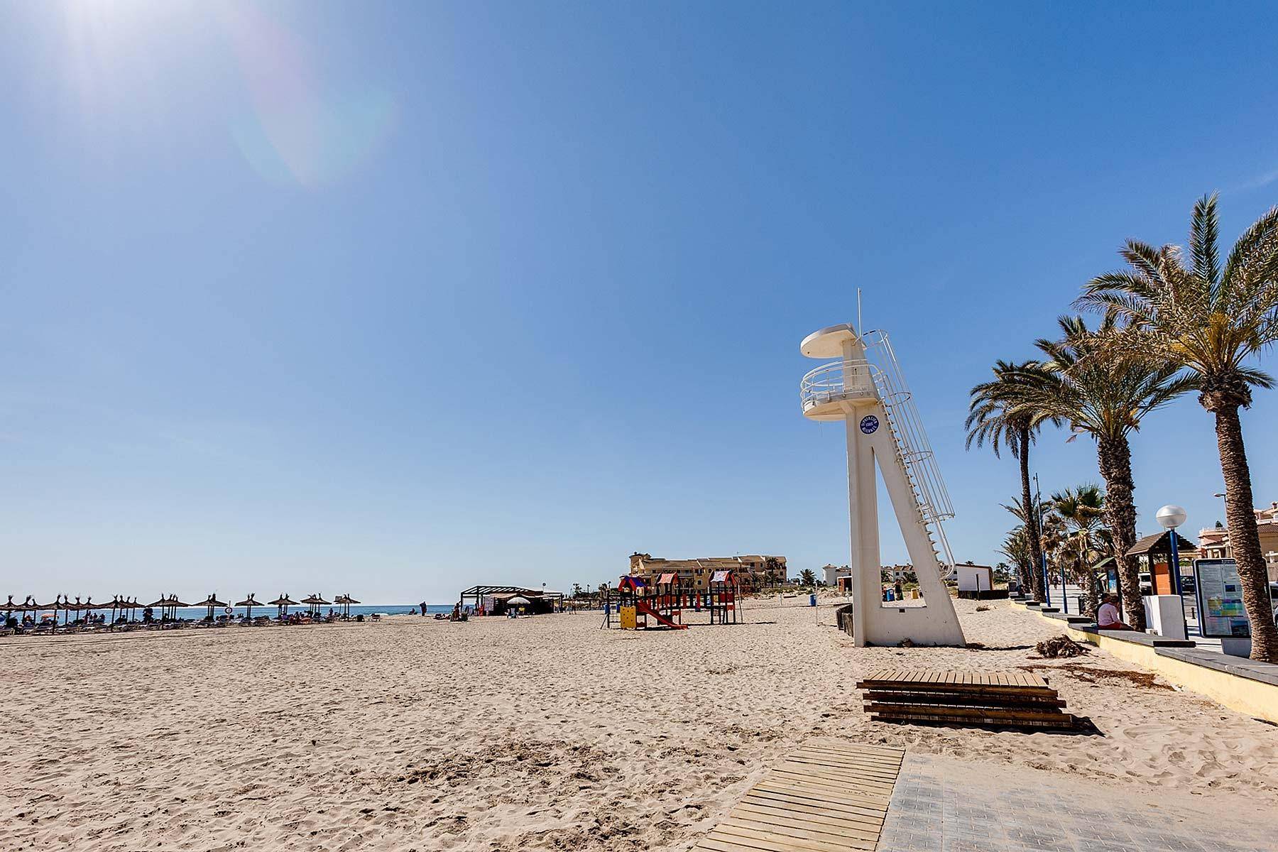 Breiter Sandstrand mit Rettungsturm, Spielplatz und Palmen unter blauem Himmel.
