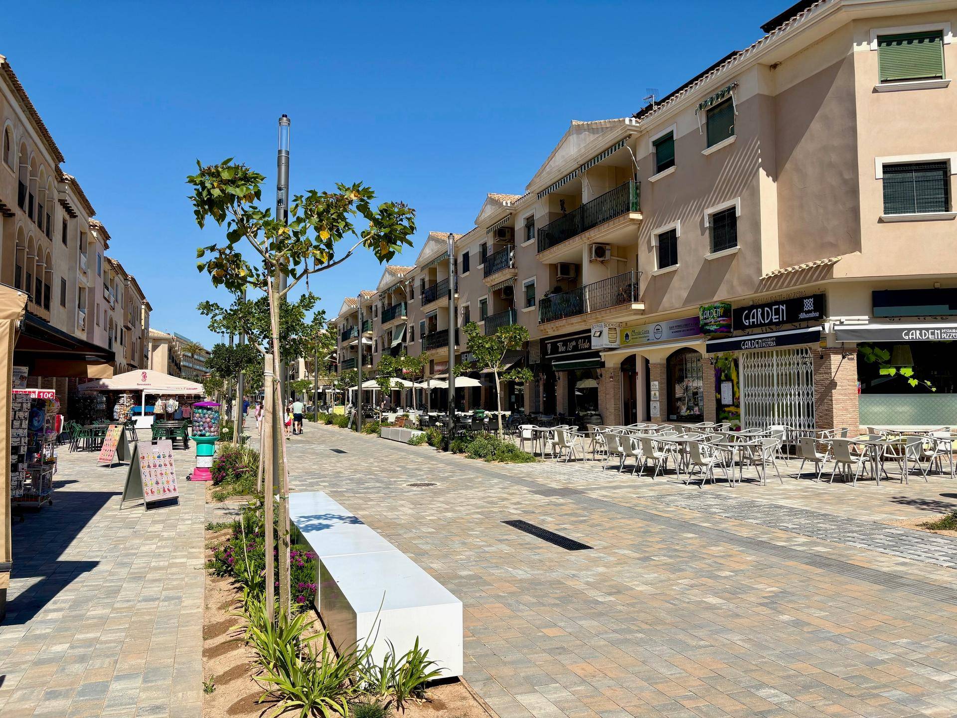 Belebte Fußgängerzone mit Geschäften, Cafés und Sitzgelegenheiten unter blauem Himmel.