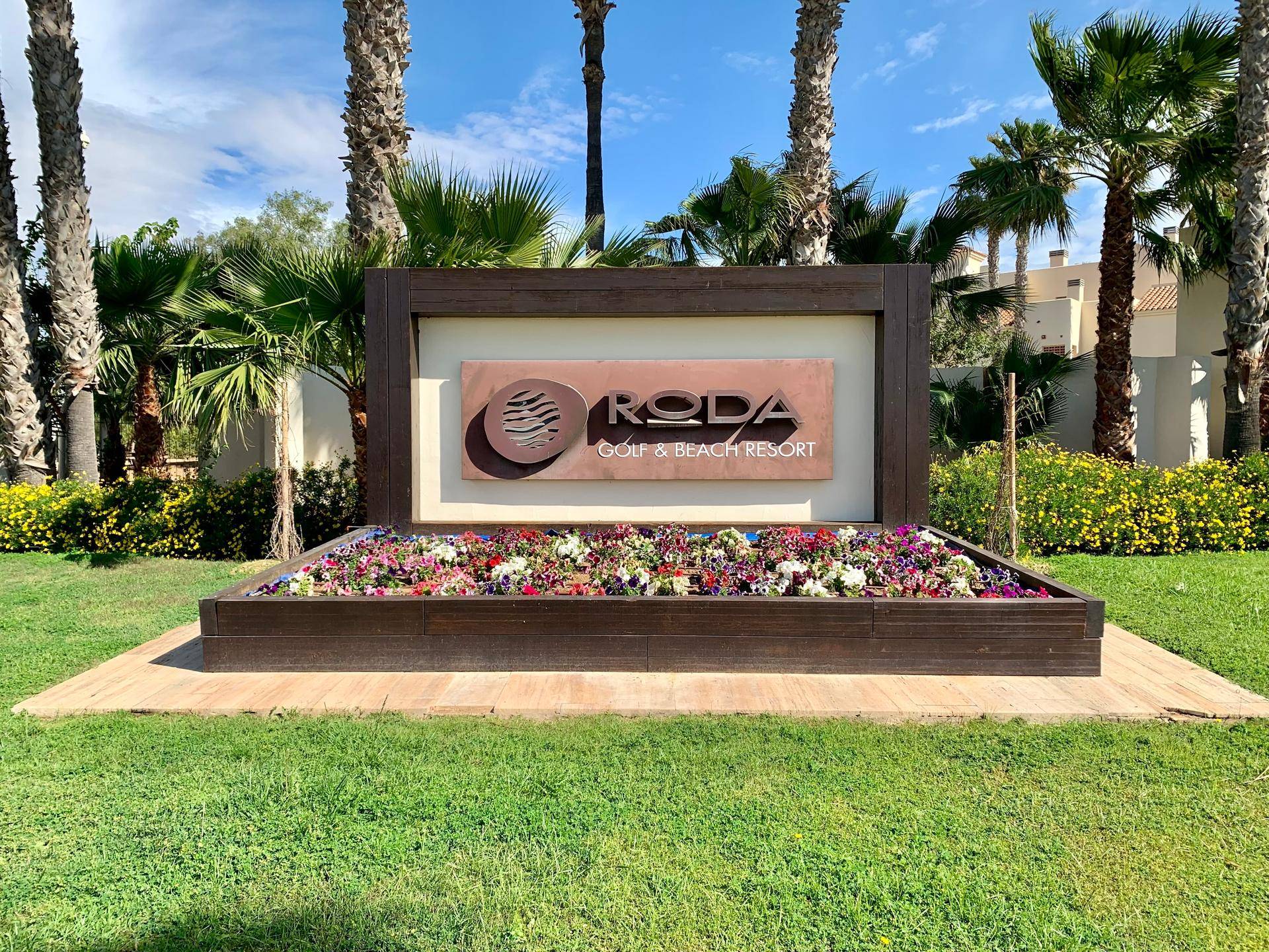 Eingangsschild des Roda Golf & Beach Resorts, umgeben von Blumenbeeten und Palmen.