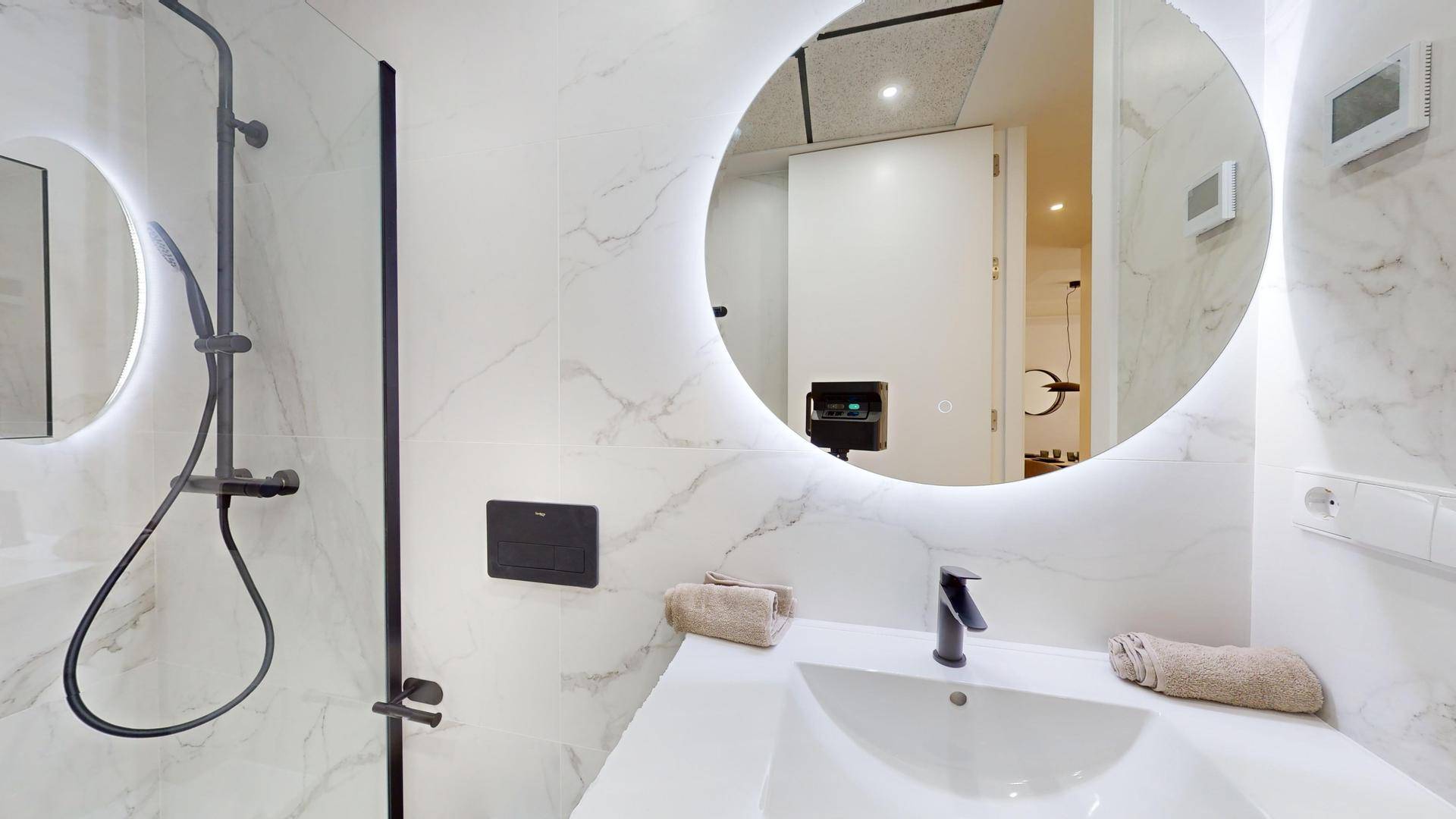 Elegantes Badezimmer mit rundem, beleuchtetem Spiegel, modernen Armaturen und Marmor-Effekt-Fliesen.