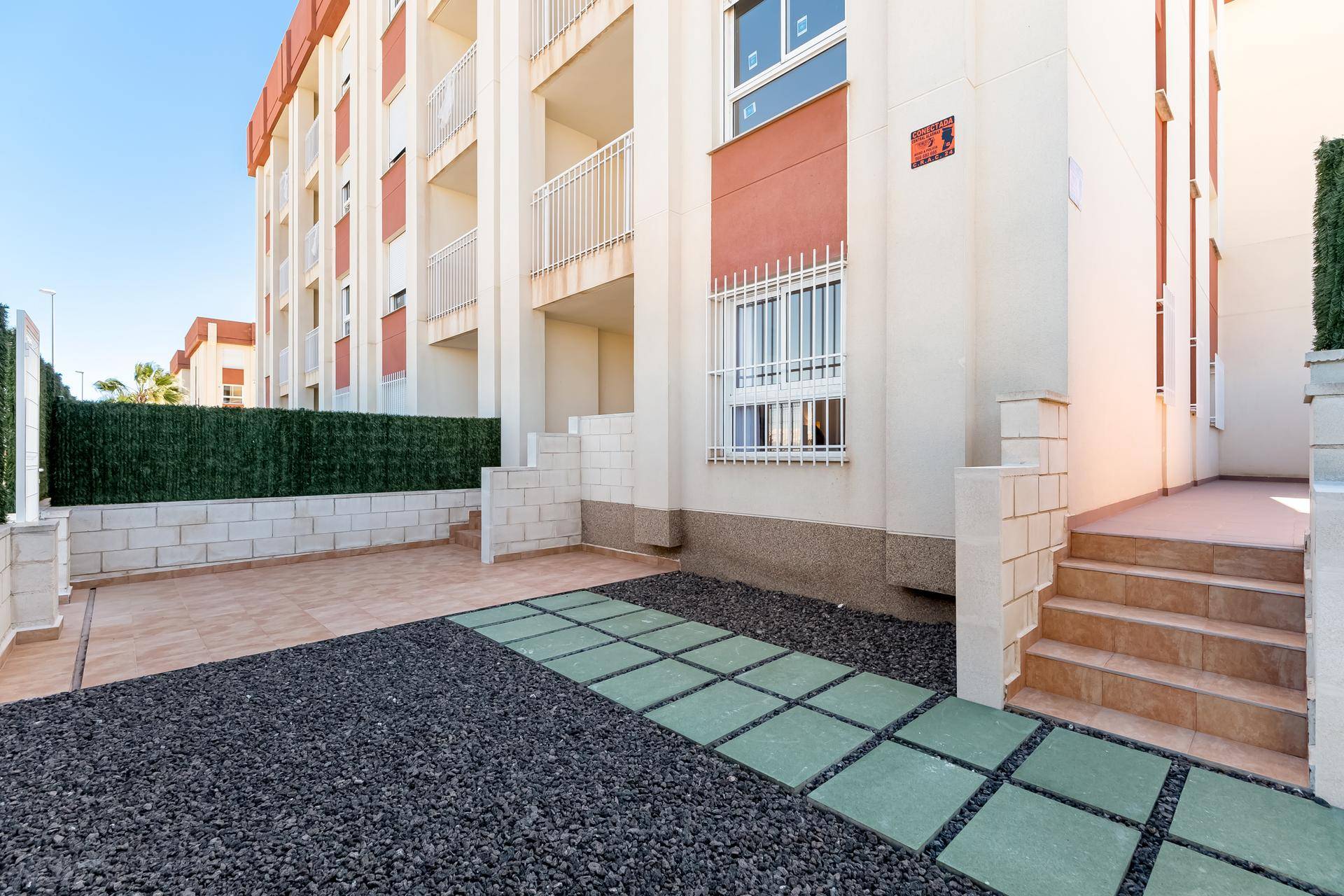 Außenbereich mit gefliester Terrasse, Zierkies und einer grünen Hecke entlang der Gebäudefassade.