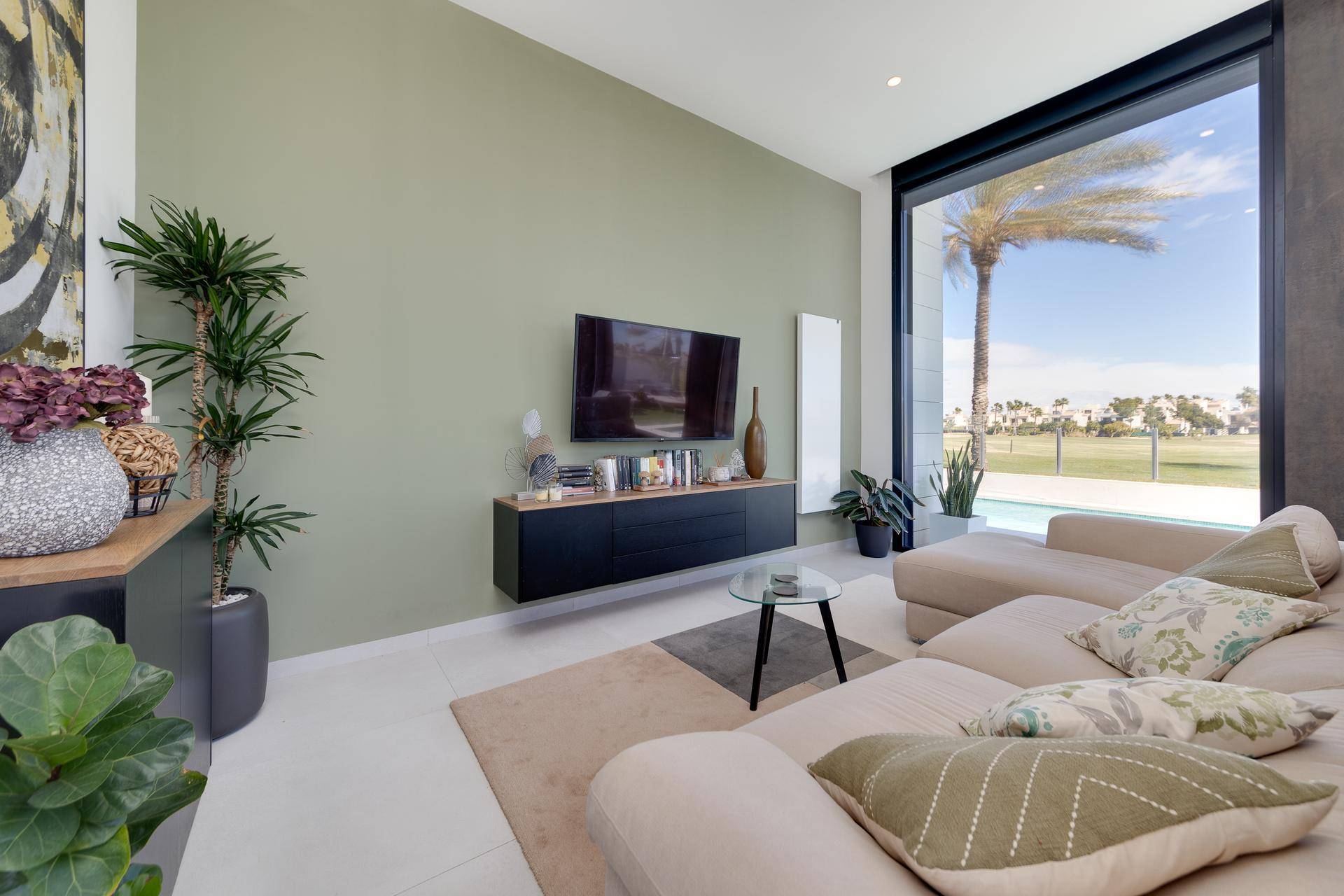 Heller Wohnbereich mit beigem Sofa, grüner Akzentwand und großen Fenstern mit Poolblick.