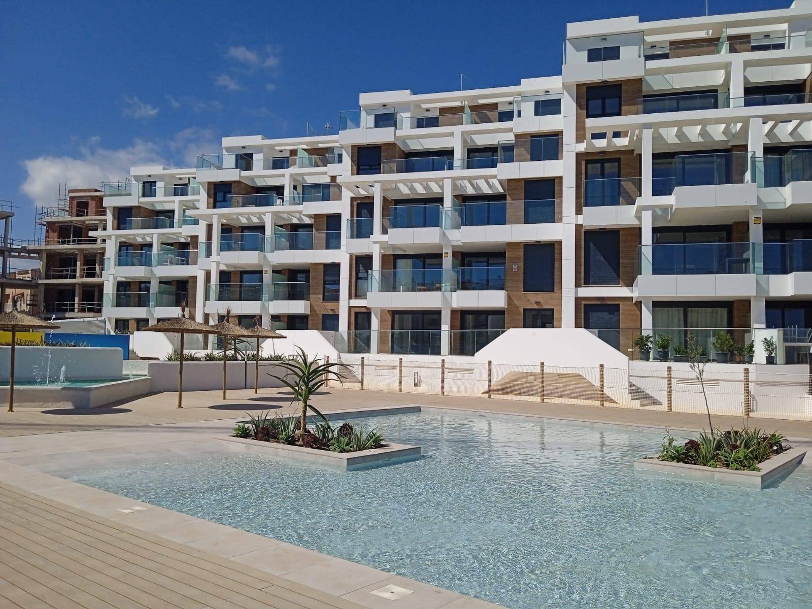 Modernes Apartmentgebäude mit weitläufigem Poolbereich, flachen Wasserzonen und Strohschirmen.