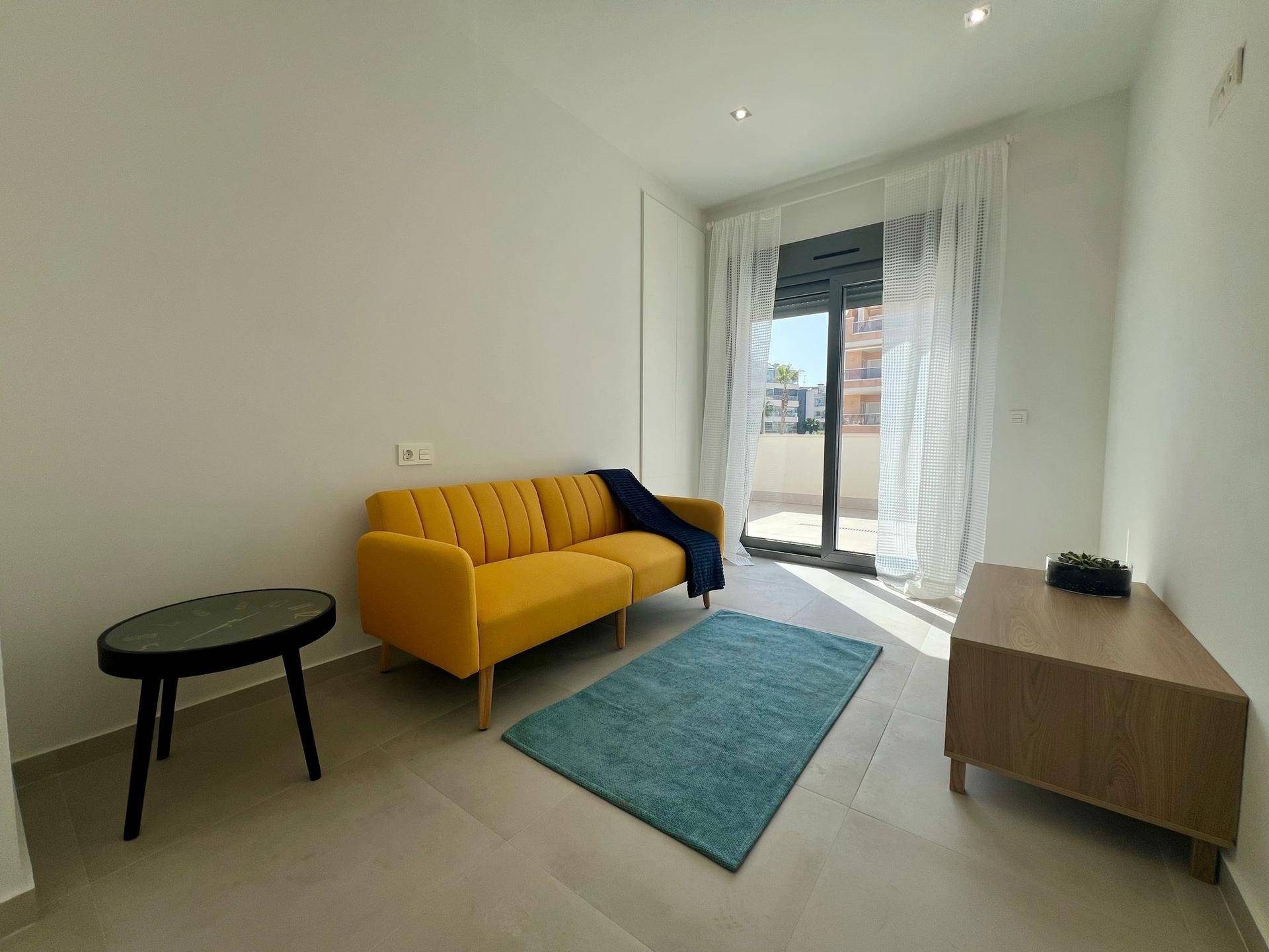 Helles Zimmer mit gelbem Sofa, Teppich und Zugang zu einem Balkon oder einer Terrasse.