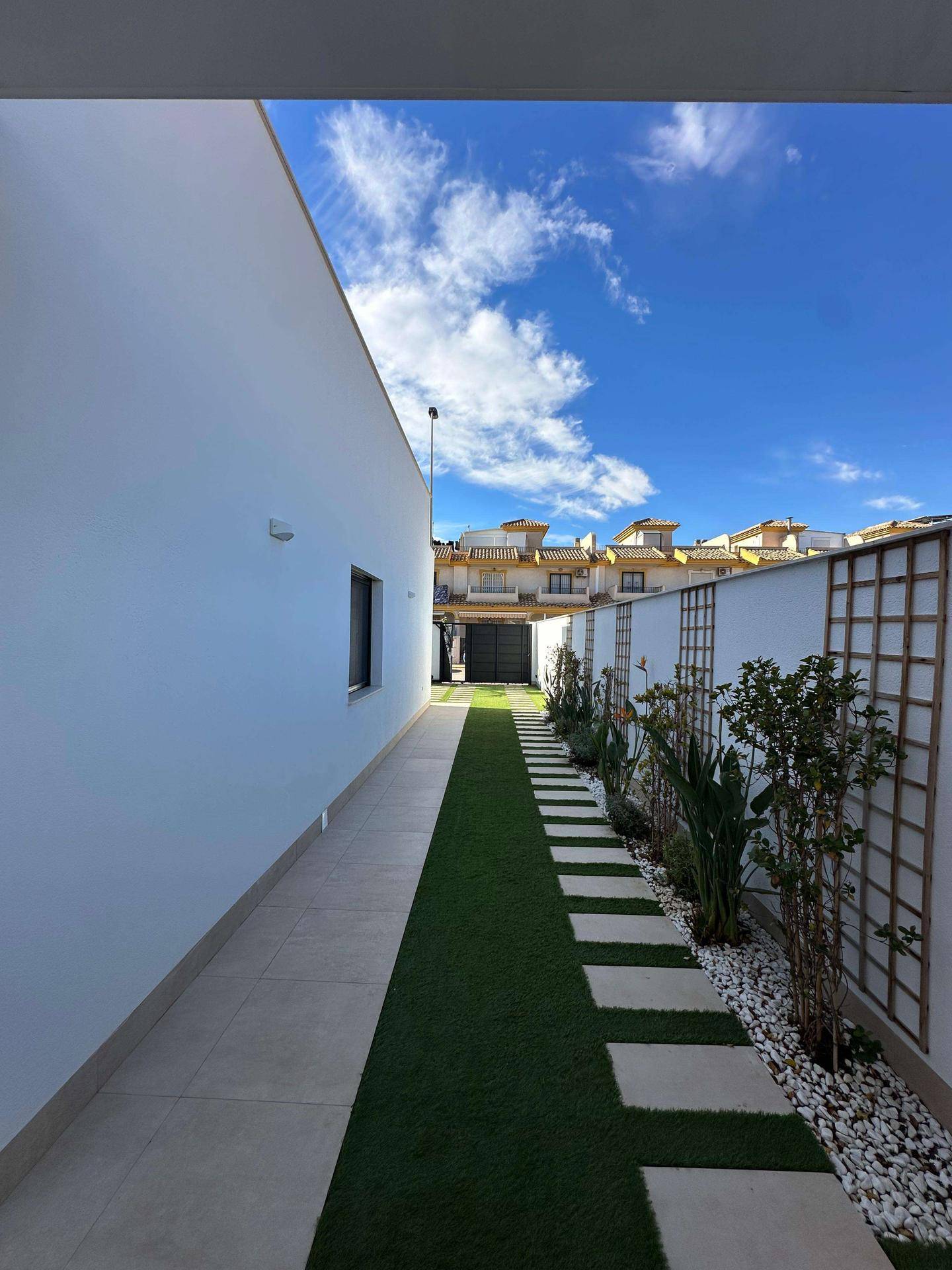 Seitlicher Gartenweg mit Kunstrasen, Trittplatten und bepflanzten Beeten entlang der Hauswand.