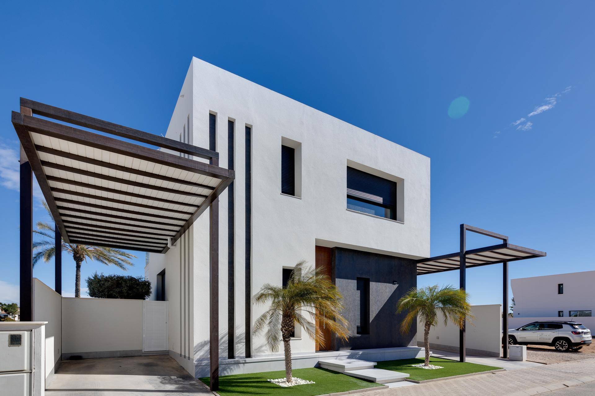 Moderne Hausfassade mit klaren Linien, Holztür und überdachtem Parkplatz unter blauem Himmel.