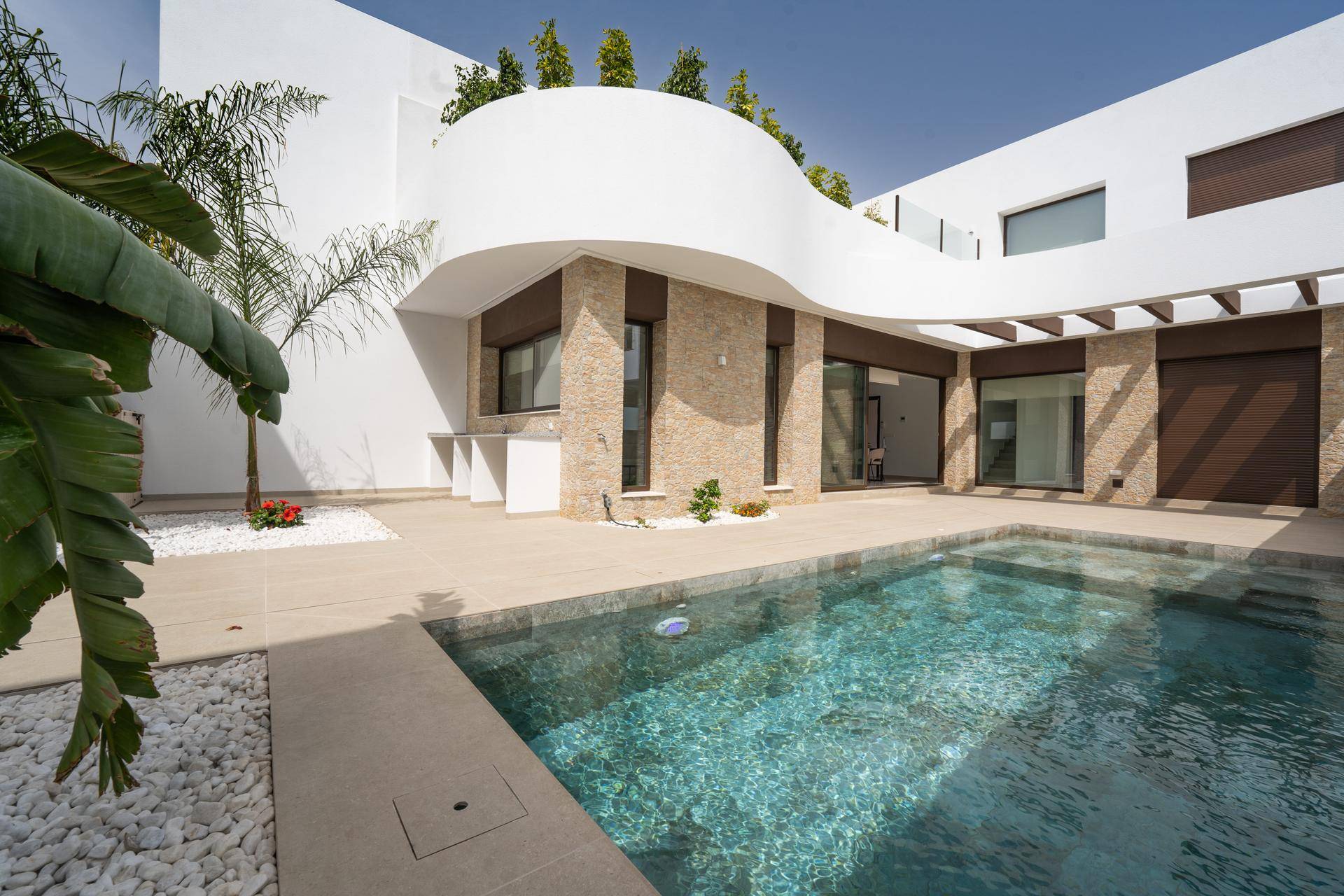 Moderne Villa bei Tageslicht mit Swimmingpool und Außenküche, umgeben von minimalistischer Landschaftsgestaltung.