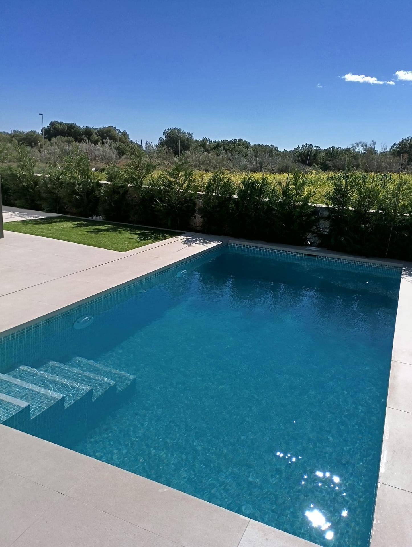 Großzügiger Pool mit breiten Einstiegsstufen und umlaufender heller Terrasse unter strahlendem Himmel.