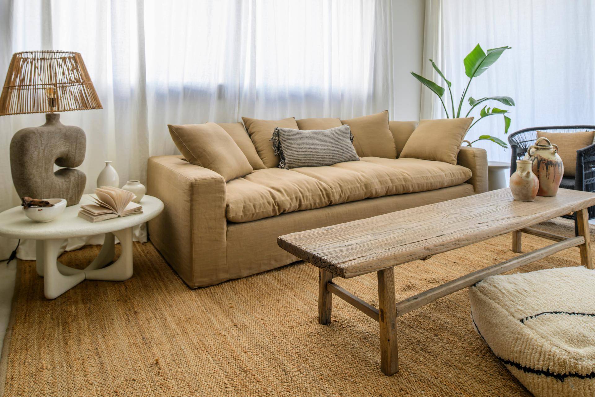 Geräumiger Wohnbereich mit beigem Sofa, Holztisch und großer Pflanze, umgeben von hellen Vorhängen.
