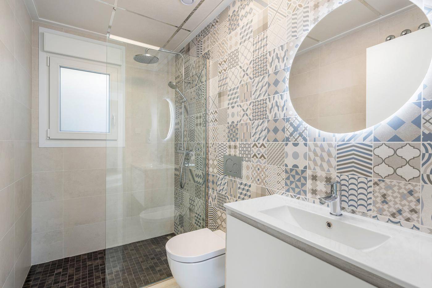 Modernes Badezimmer mit gemusterten Fliesen, begehbarer Dusche und beleuchtetem Rundspiegel.