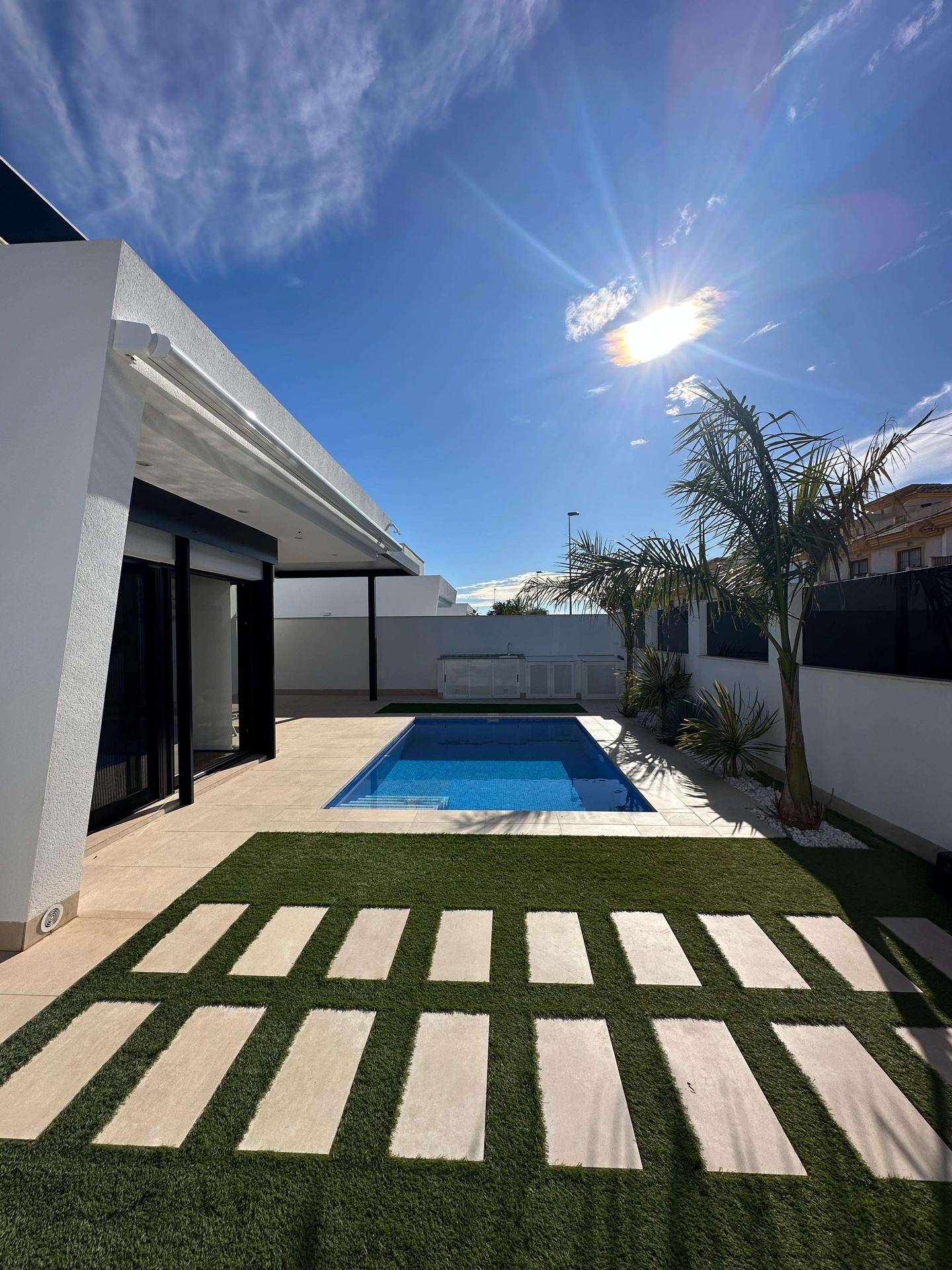 Sonniger Außenbereich mit Pool, Kunstrasen und Trittplatten, umgeben von Palmen.