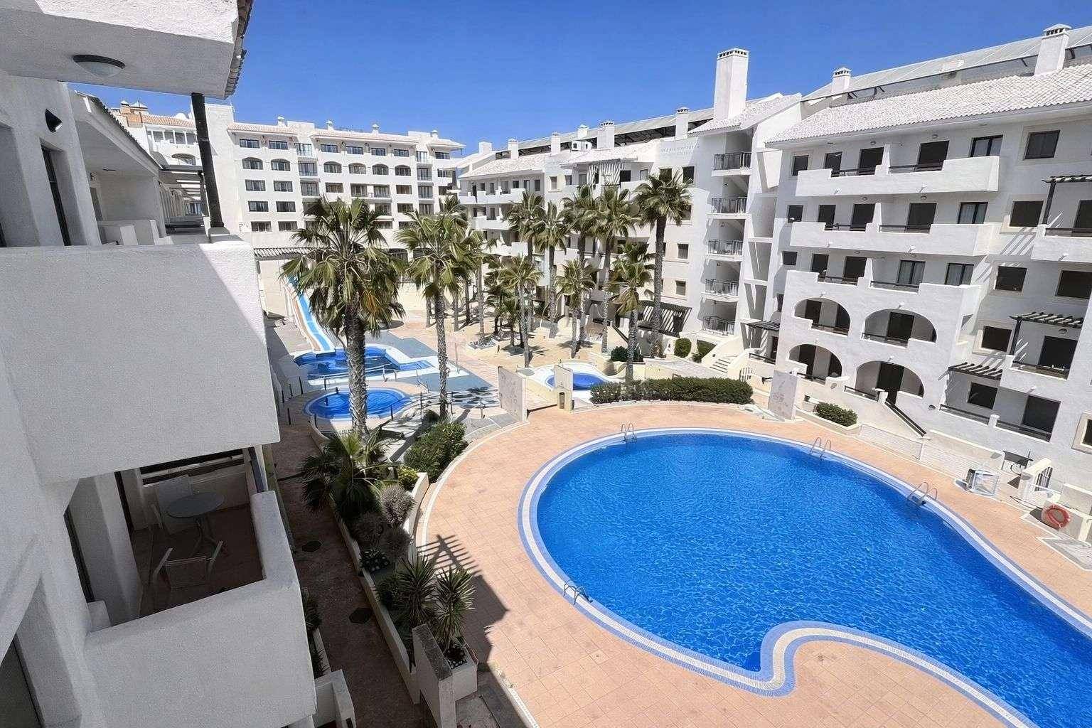 Innenhof eines Apartmentkomplexes mit großem Pool, Palmen und einer Wasserrutsche im Hintergrund.