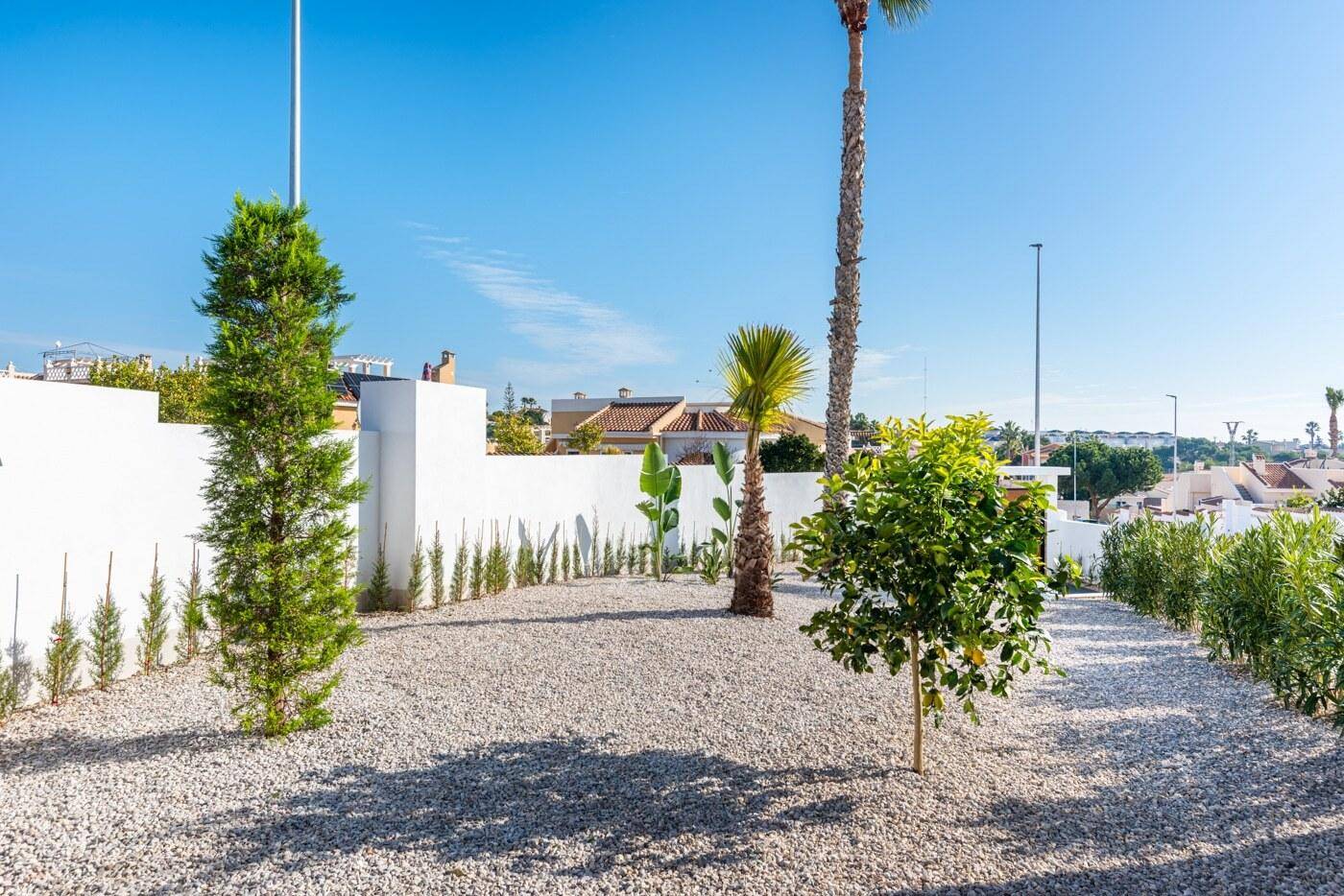Mediterraner Gartenbereich mit Kies, Palmen und jungen Bäumen unter blauem Himmel.