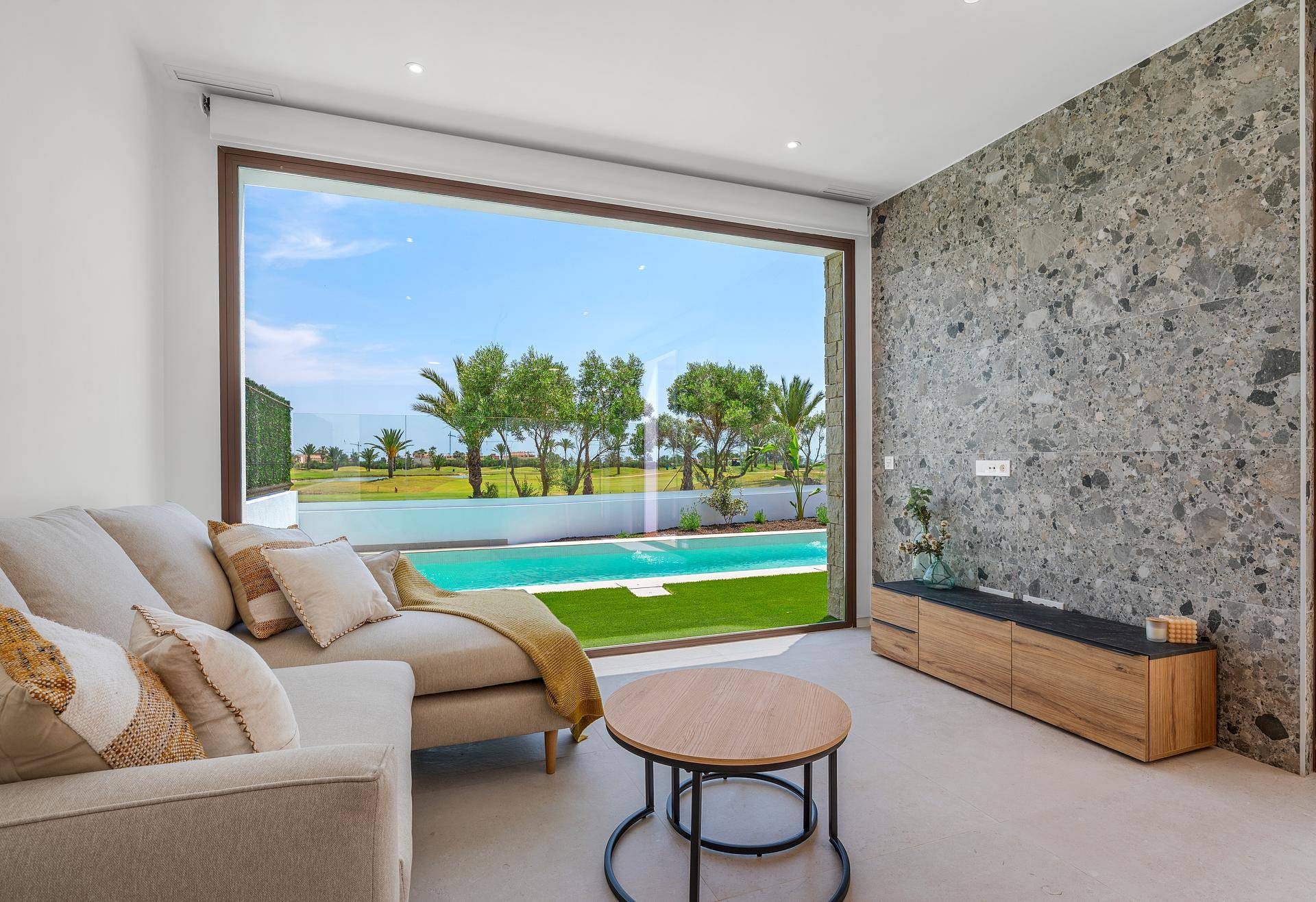Gemütliches Wohnzimmer mit Sofa, Couchtisch und raumhohen Fenstern mit Poolblick.