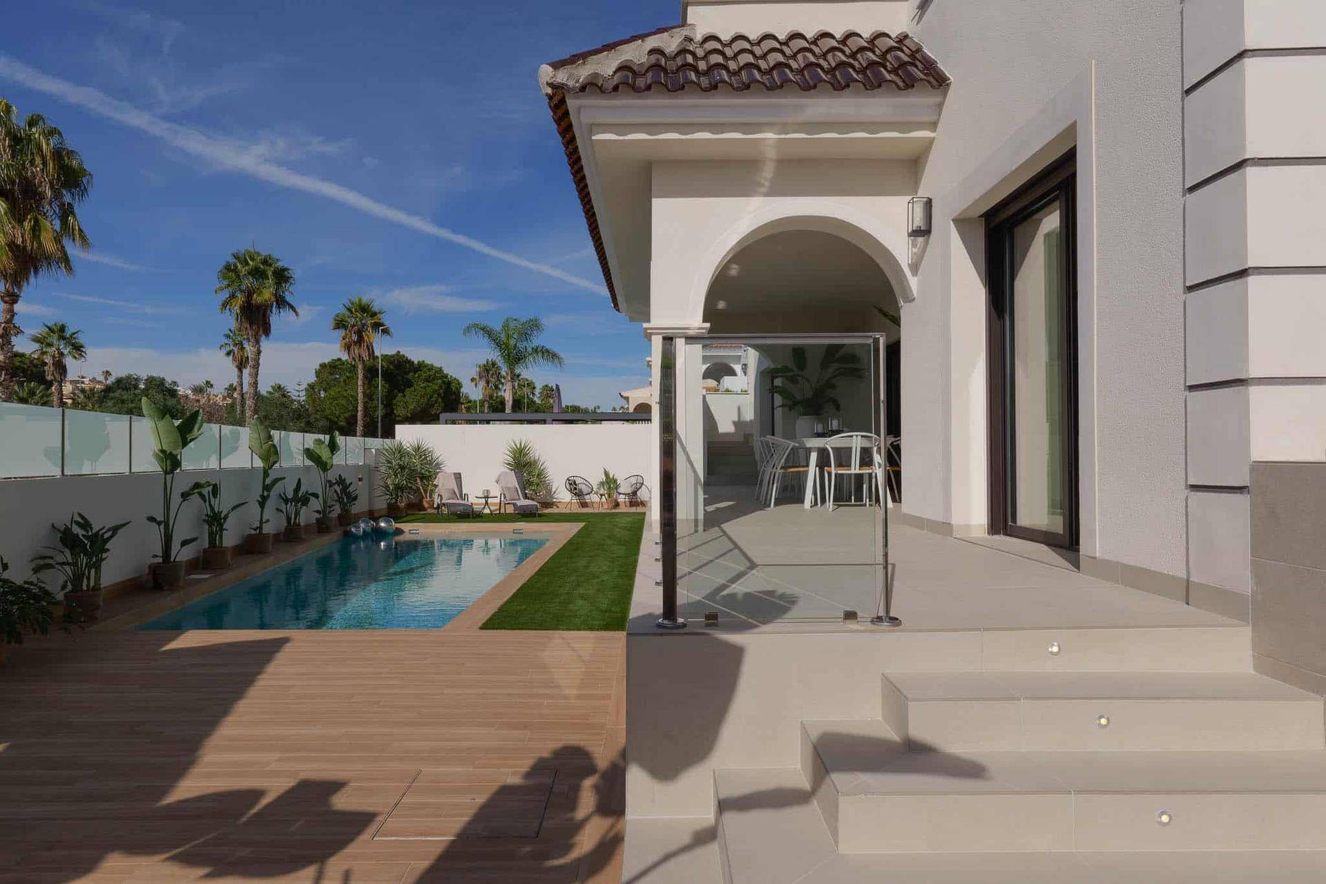 Elegante Hausfassade mit Zugang zum Poolbereich und einer modernen Glasbalustrade unter blauem Himmel.