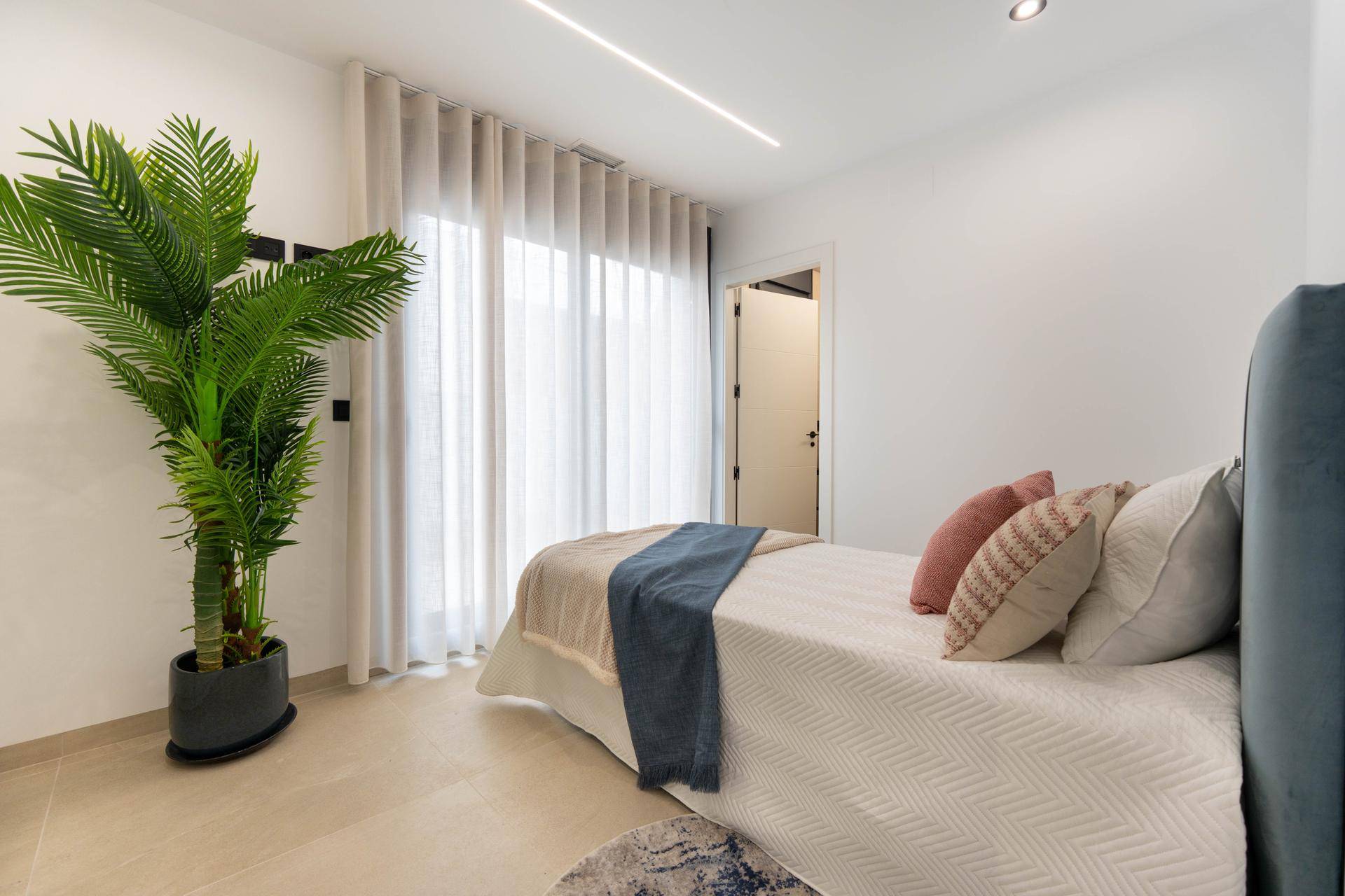 Gemütliches Schlafzimmer mit großen Fenstern, Vorhängen und einer dekorativen Palme.