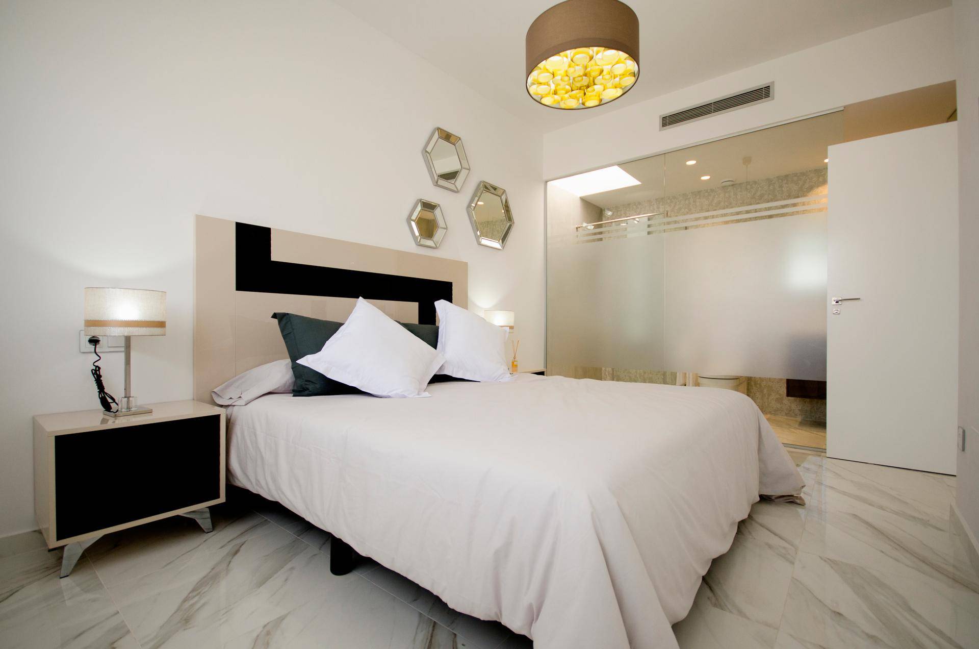 Helles Schlafzimmer mit Doppelbett, Nachttisch und offenem Zugang zum modernen Bad.