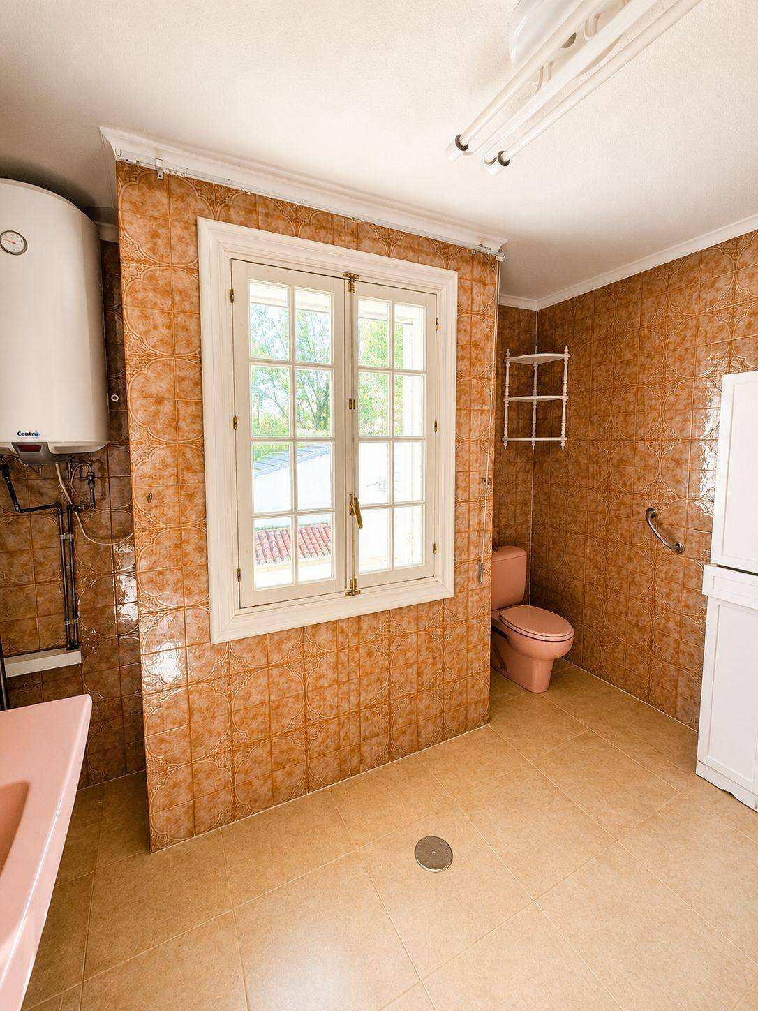 Badezimmer mit beigen Fliesen, rosa Toilette und Waschbecken sowie einem Fenster für Tageslicht.