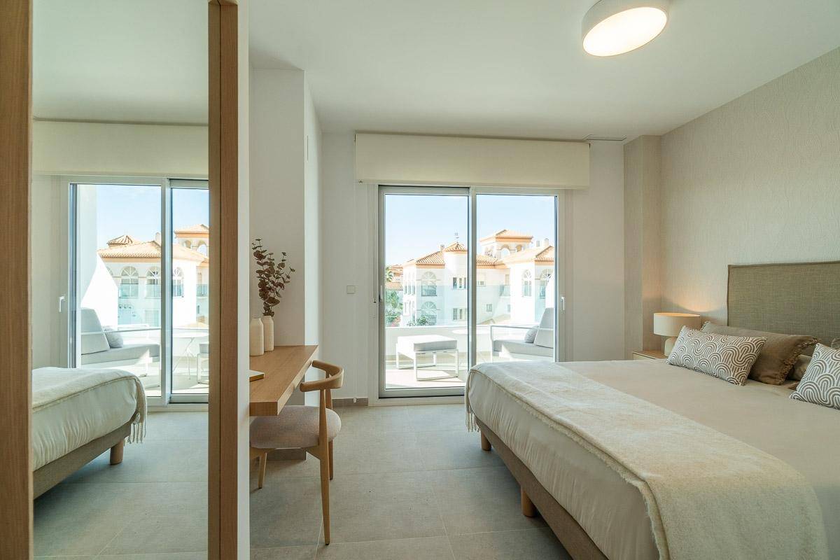 Schlafzimmer mit Doppelbett, Schreibtisch und großen Schiebetüren, die zu einem sonnigen Balkon führen.