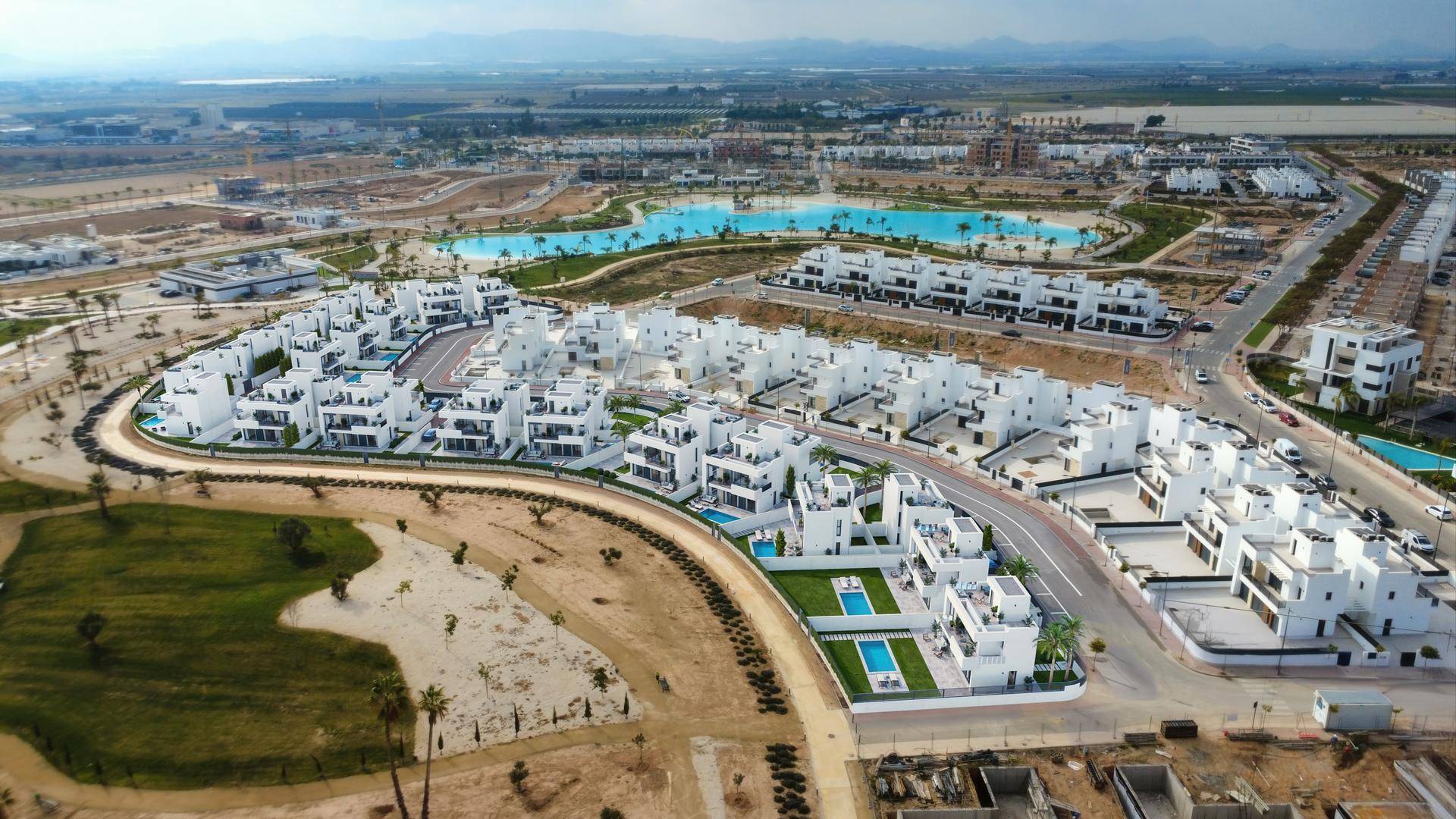 Luftaufnahme einer modernen Wohnanlage mit zahlreichen Villen, Pools und einem großen See in der Ferne.